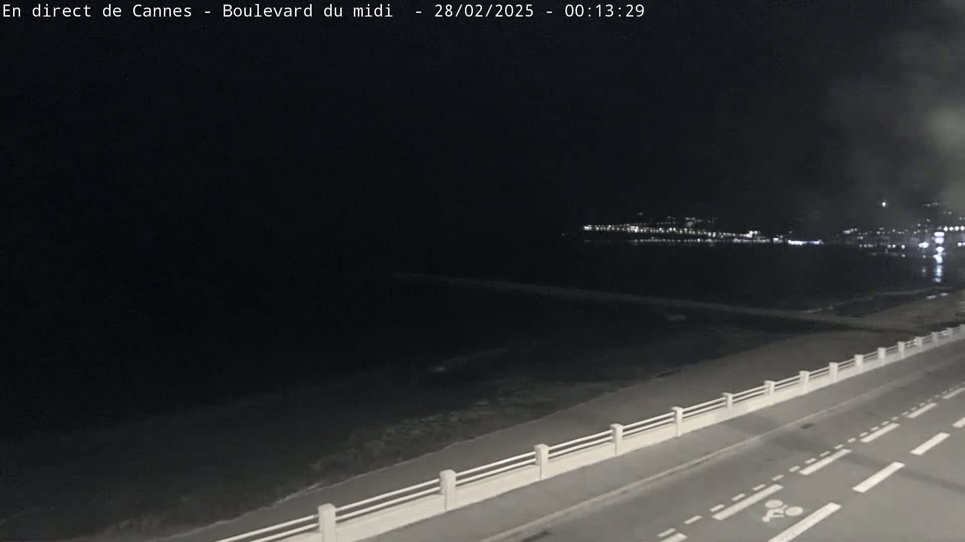 A nighttime, seaside view shows a dark road with a low white railing next to a dark beach and a faintly lit cityscape across the water.