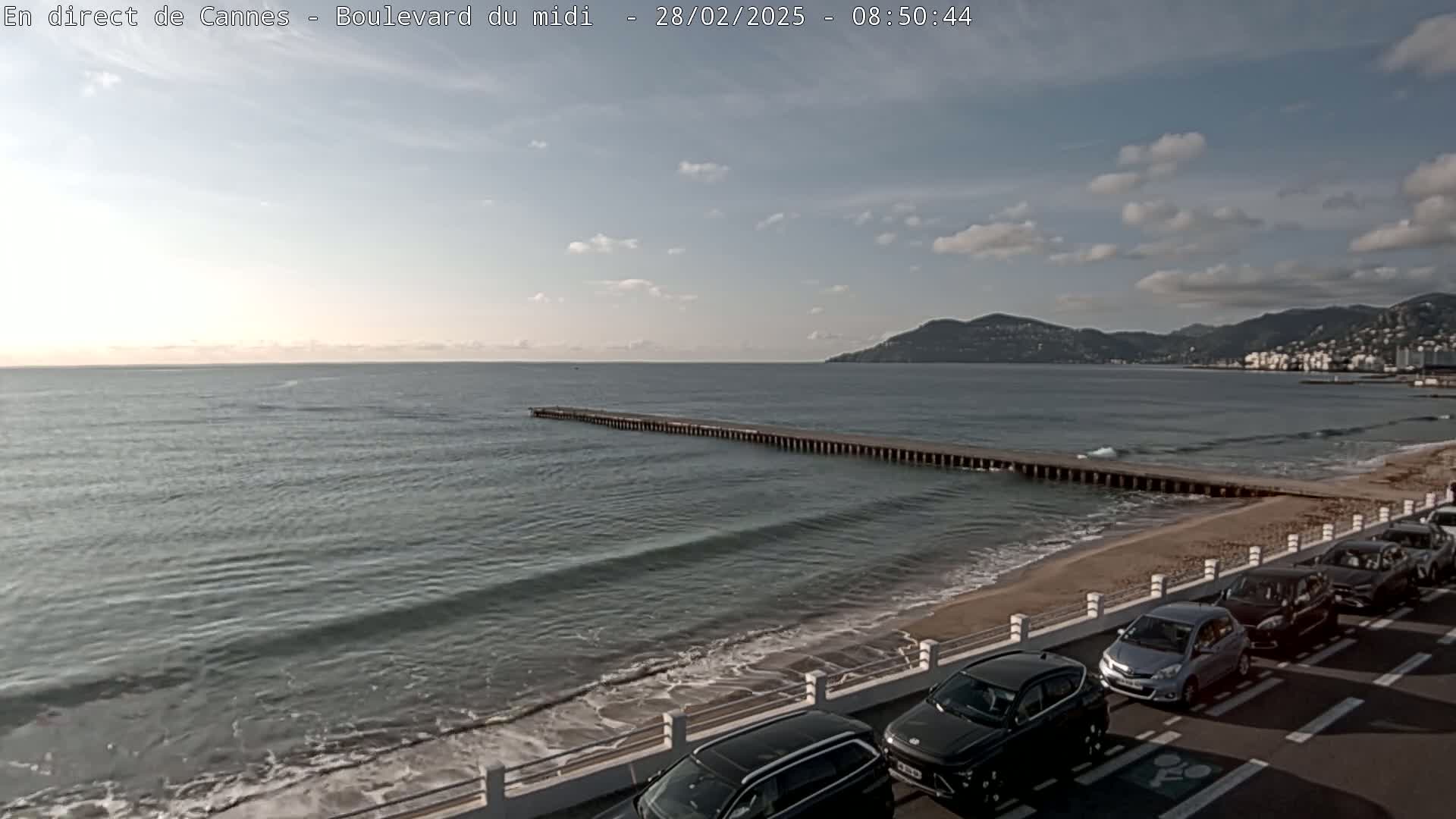 A mostly sunny day shows a long pier extending into a calm sea with gentle waves lapping a sandy beach where several cars are parked along a roadside.