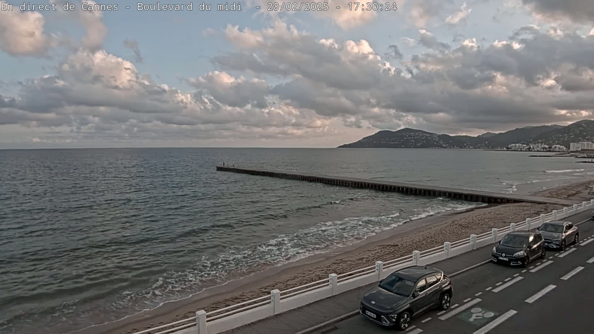 A cloudy day at a beach shows a long pier extending into calm, gray water with several cars parked along the road next to the sand.