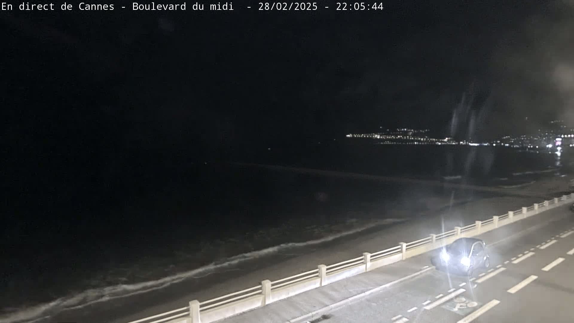 A dark nighttime scene shows a car driving along a coastal road next to a beach with gentle waves under a clear, dark sky.