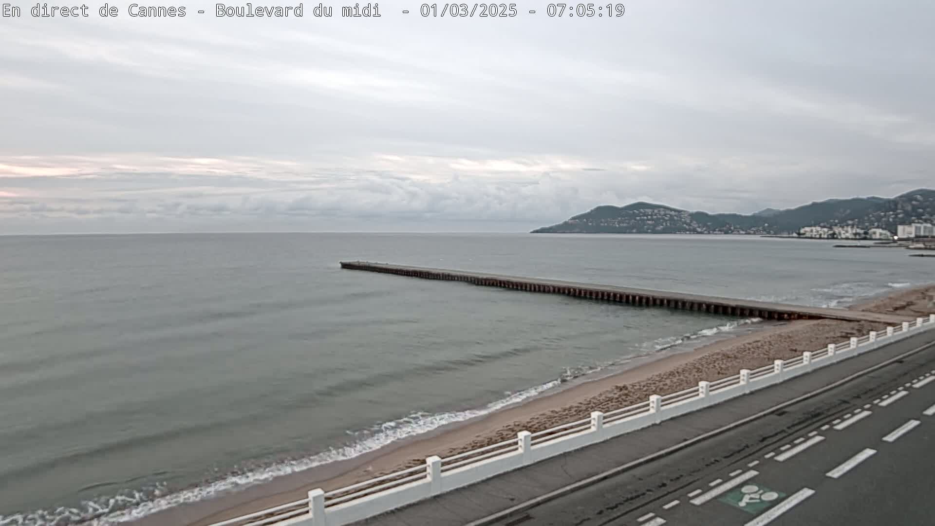 A long pier extends into a calm, gray sea under a cloudy sky, next to a sandy beach and road.