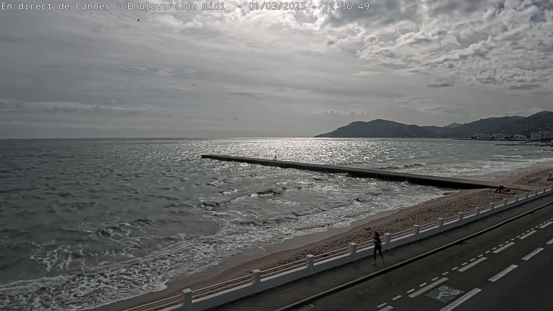 A person jogs along a road next to a beach with moderate waves under a mostly cloudy sky, a long jetty extending into the sea, and mountains visible in the distance.