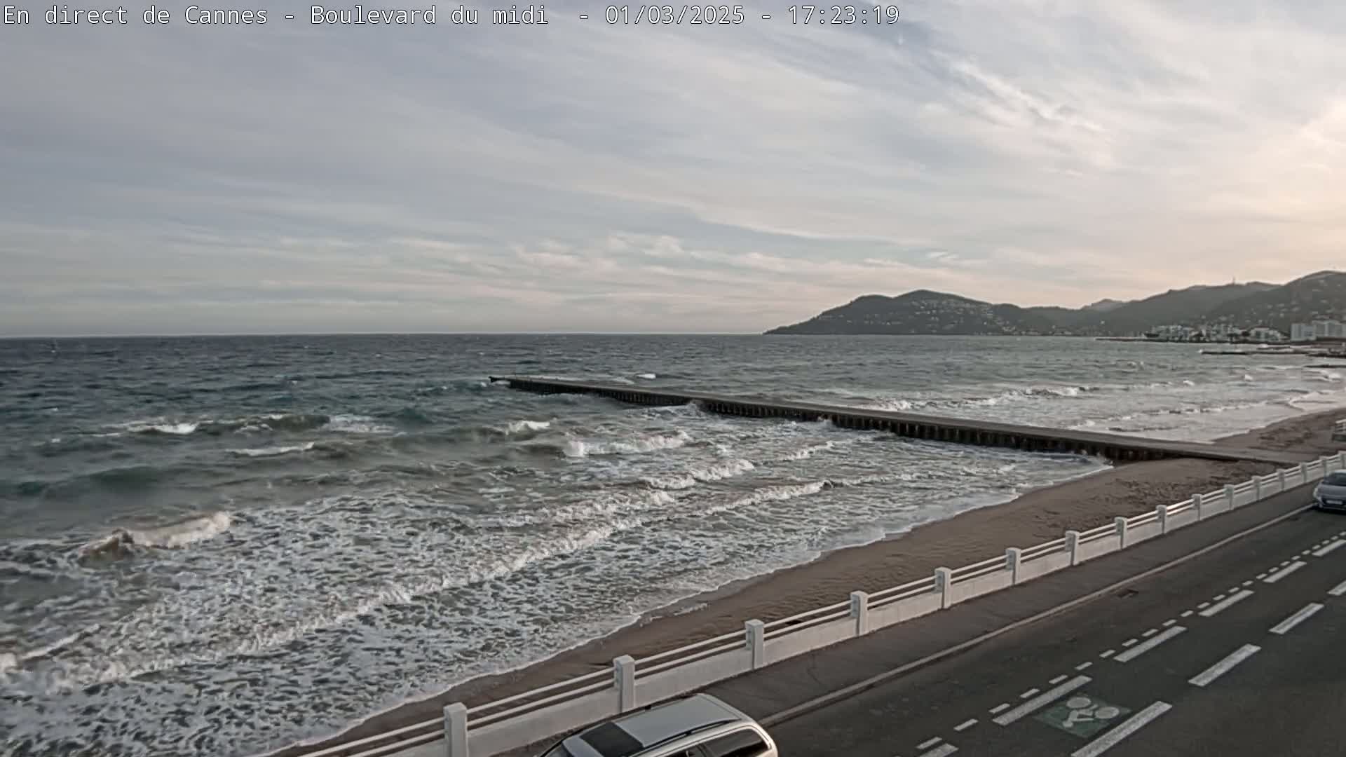 A partly cloudy day shows a beach with moderate waves crashing onto the shore, a long pier extending into the water, and a road with cars alongside a white fence.