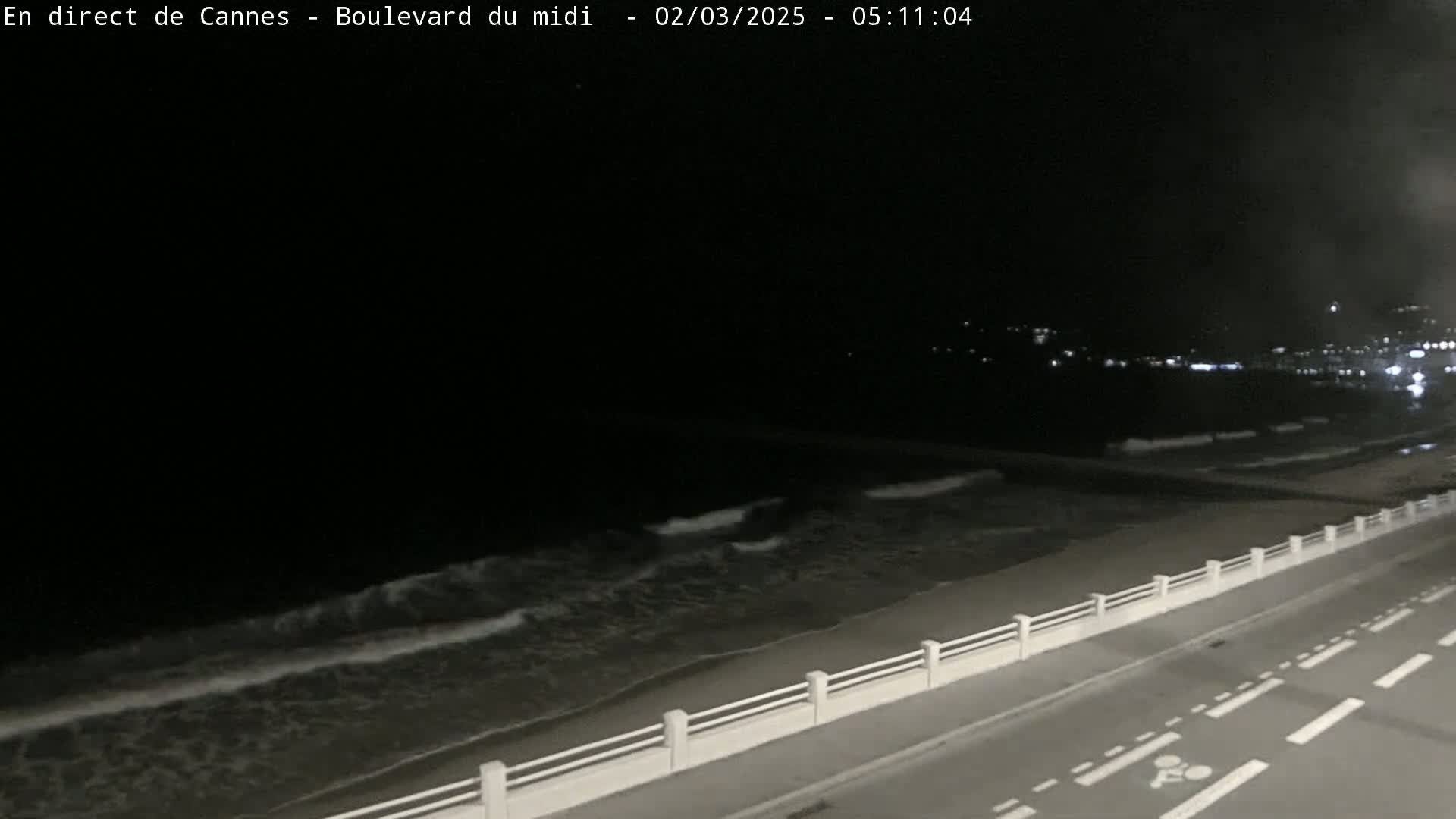 A nighttime view of a beach with gentle waves, a road with bike lanes, and a low white wall separating the road from the sand.