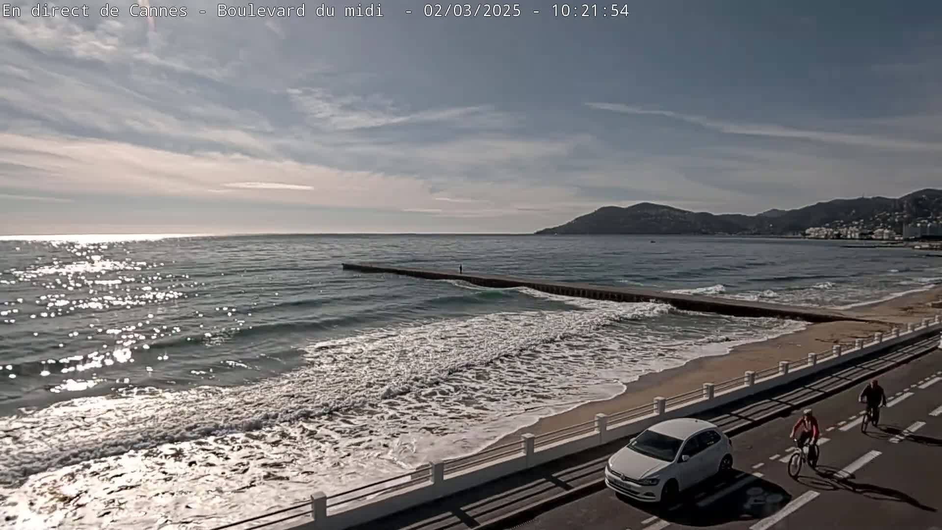 A mostly sunny day shows a beach with moderate waves, a long jetty, a car and two cyclists on a coastal road, and mountains in the distance.