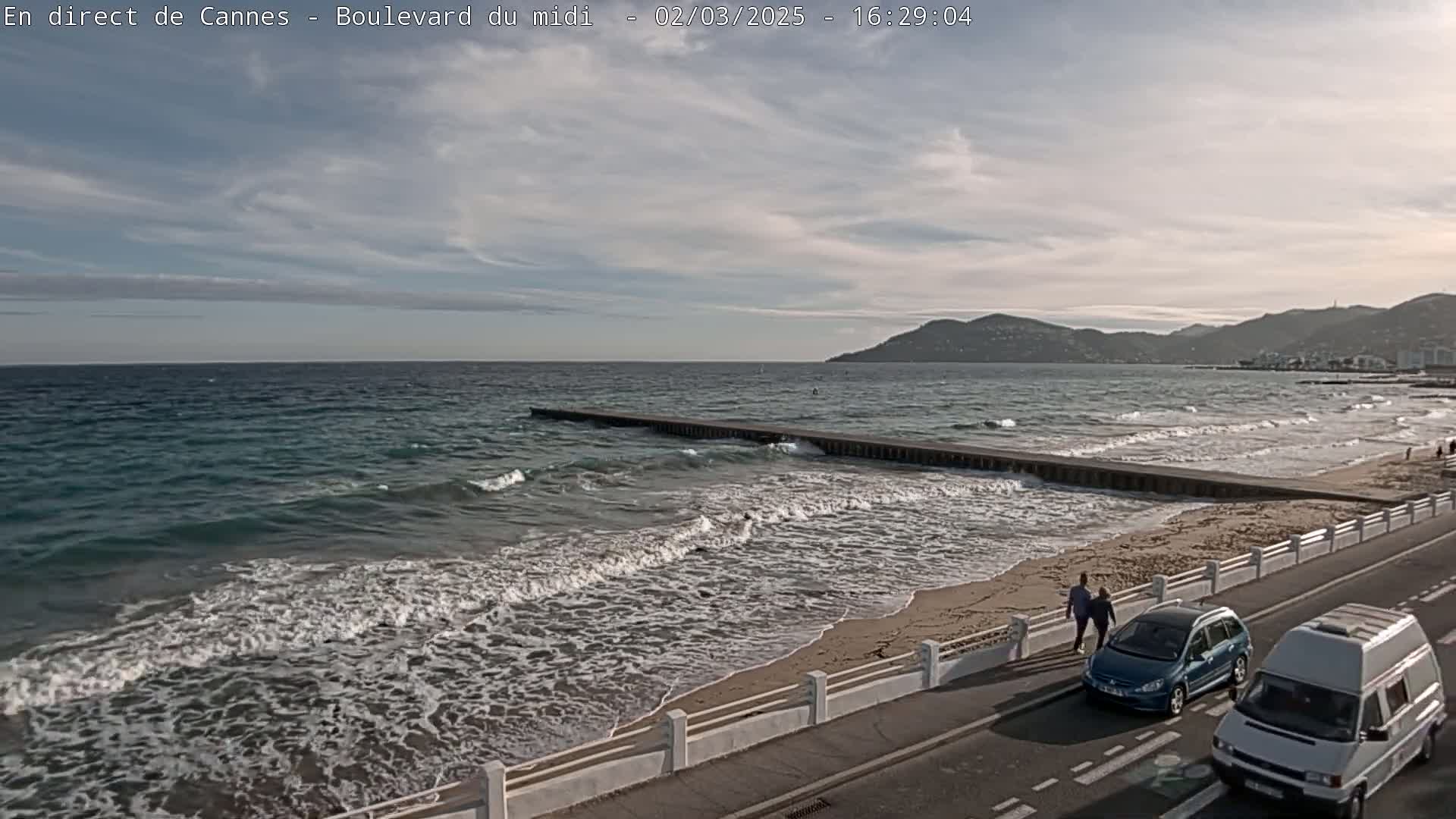 A partly cloudy day shows a beach with moderate waves breaking on the shore, a road with cars and people walking beside it, and mountains in the distance.