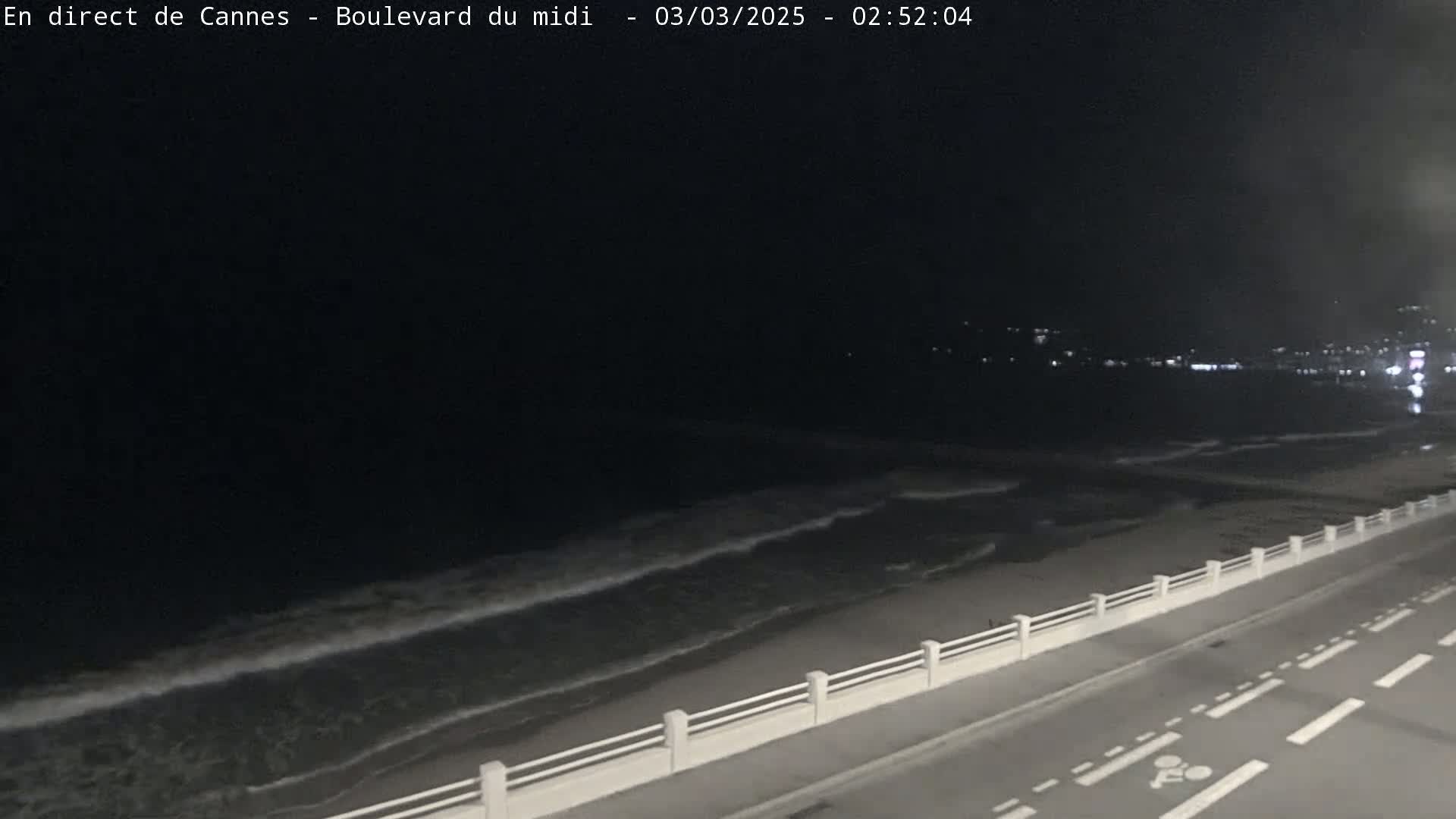A nighttime, coastal view shows dark ocean waves lapping a sandy shore next to a road with a low white railing.