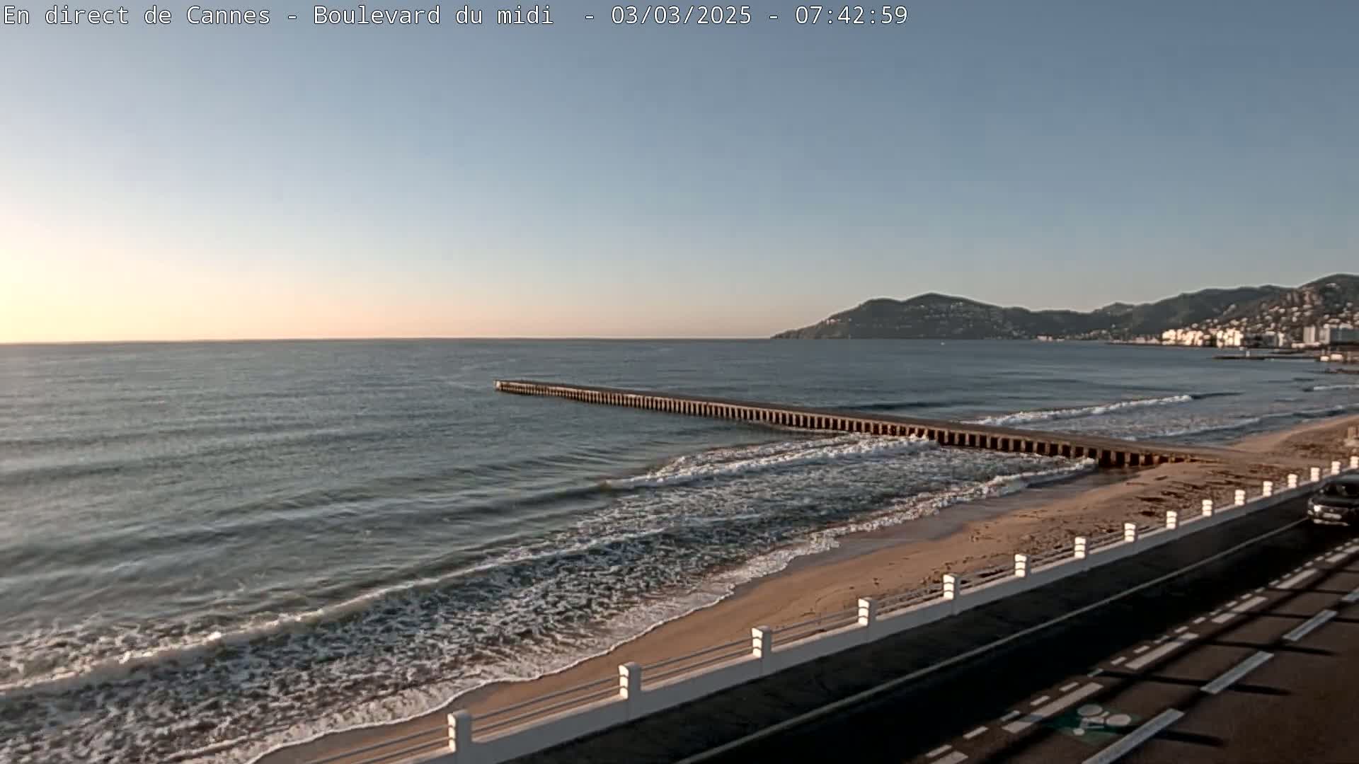 A long pier extends into a calm sea with small waves lapping a sandy beach under a clear, light blue sky.