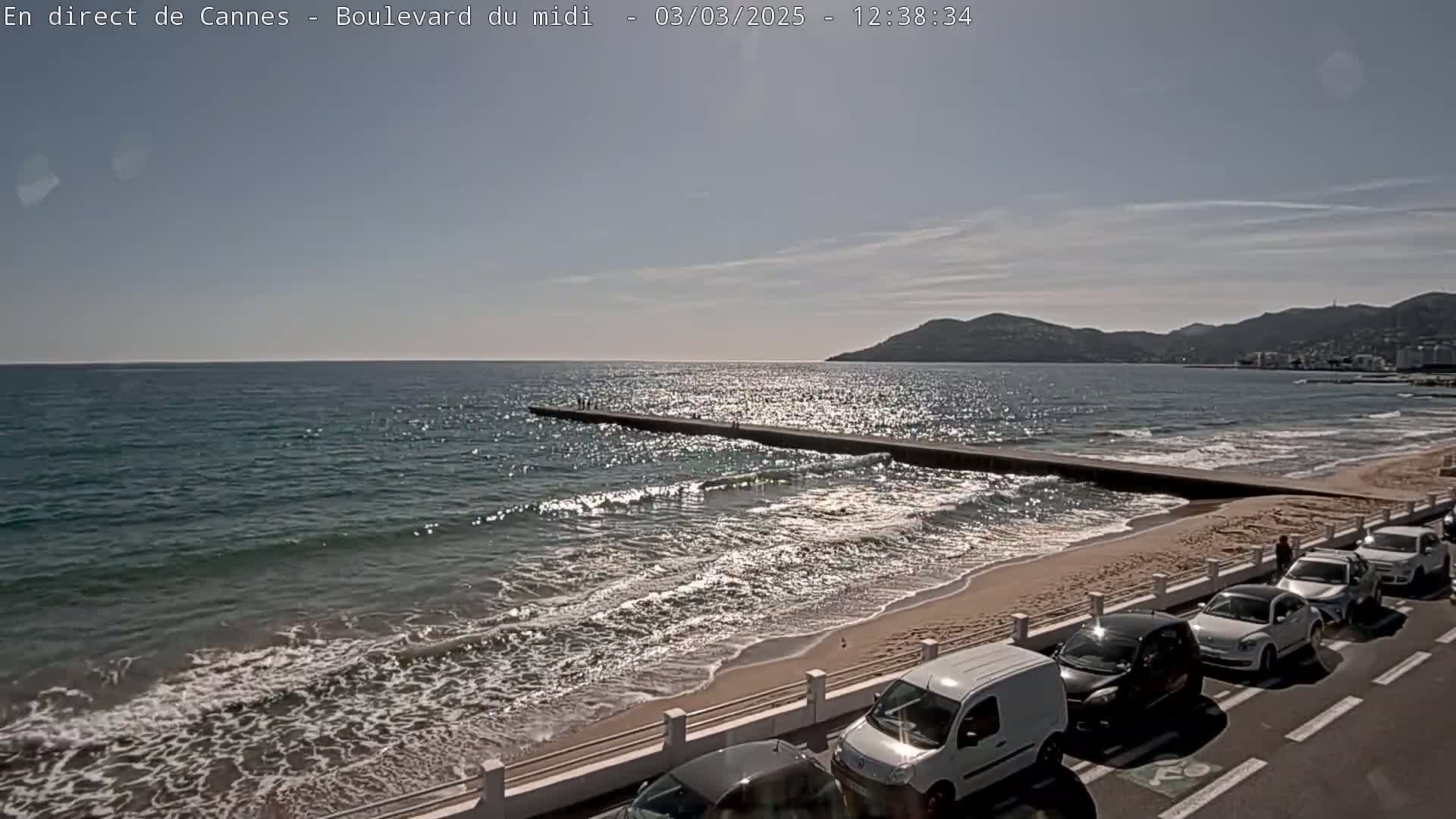 A sunny day shows a sandy beach with gentle waves lapping the shore, a long jetty extending into the sea, and cars parked along the road next to the beach.