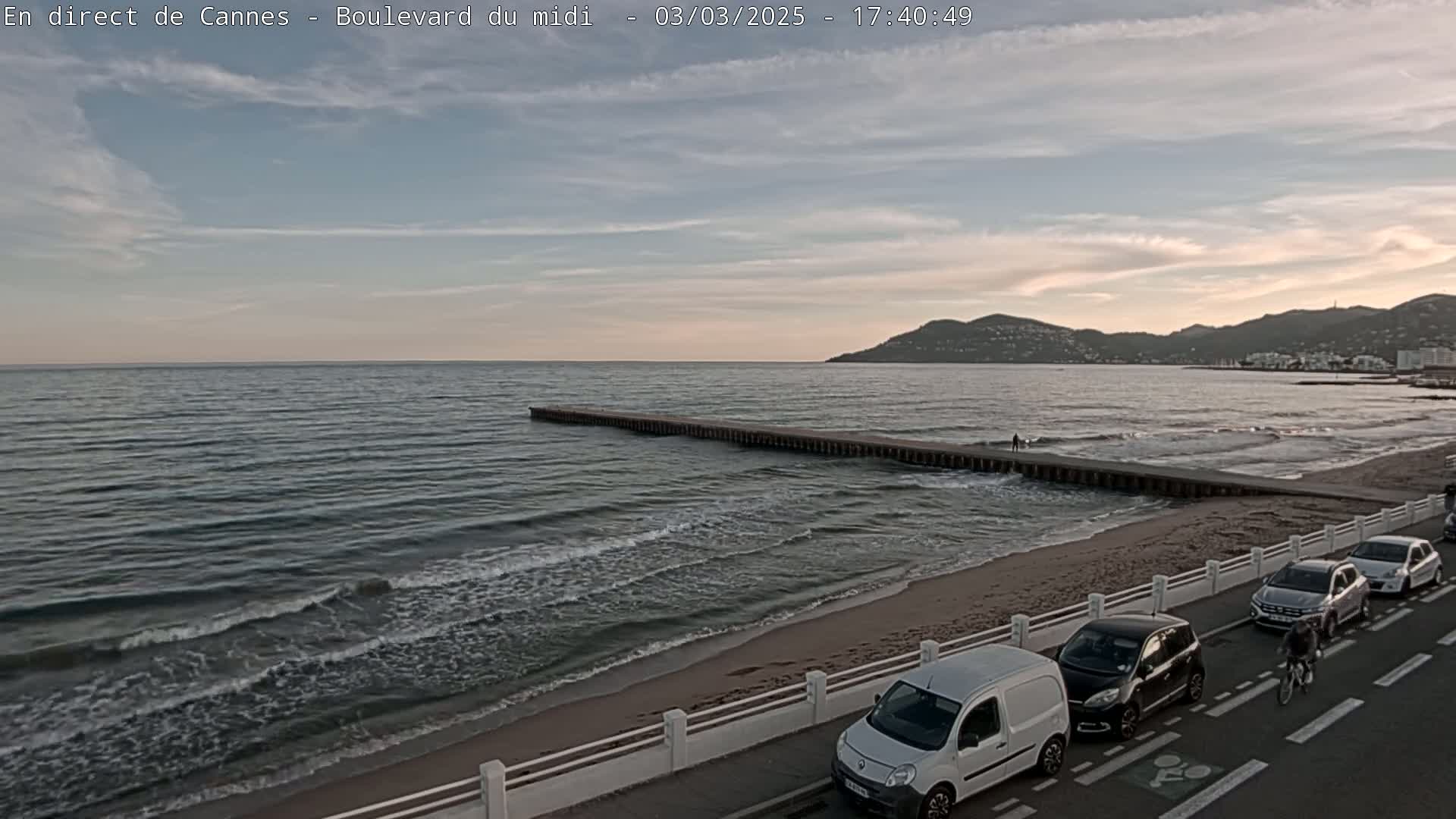 A sandy beach with gentle waves and a long pier extends into a calm sea under a partly cloudy, dusky sky, with cars parked along a road beside it and a cyclist riding by.