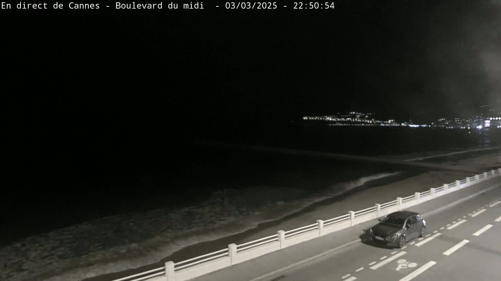 A dark gray car is parked on a road next to a beach at night, with gentle waves lapping the shore under a clear, dark sky.