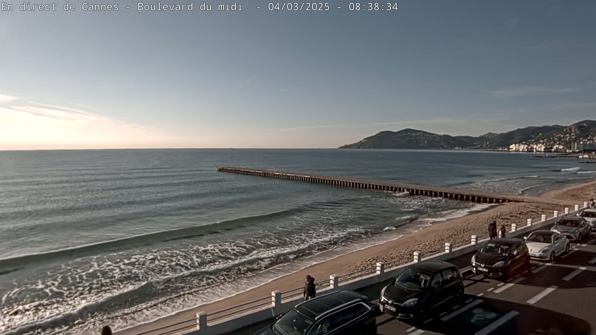 A sandy beach with gentle waves laps at the shore under a clear, bright sky, with cars parked along a road overlooking a long pier extending into the sea and distant hills in the background.