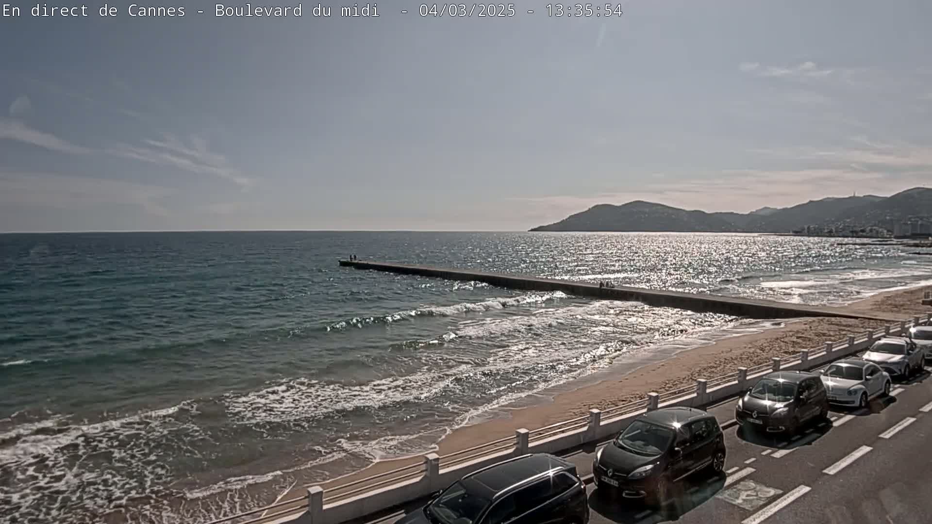 A sandy beach with gentle waves and a long pier extends into a calm, sunlit sea under a mostly clear sky, with cars parked along a road bordering the beach.