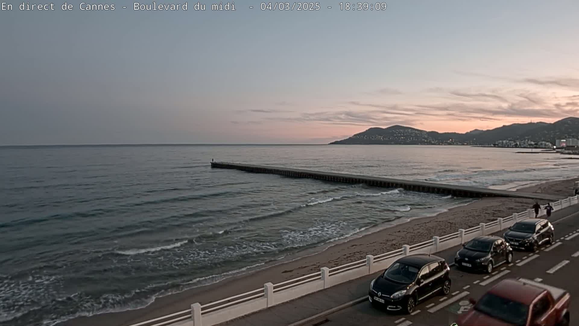A long jetty extends into a calm sea under a soft, dusky sky as several cars park along a beachside road.