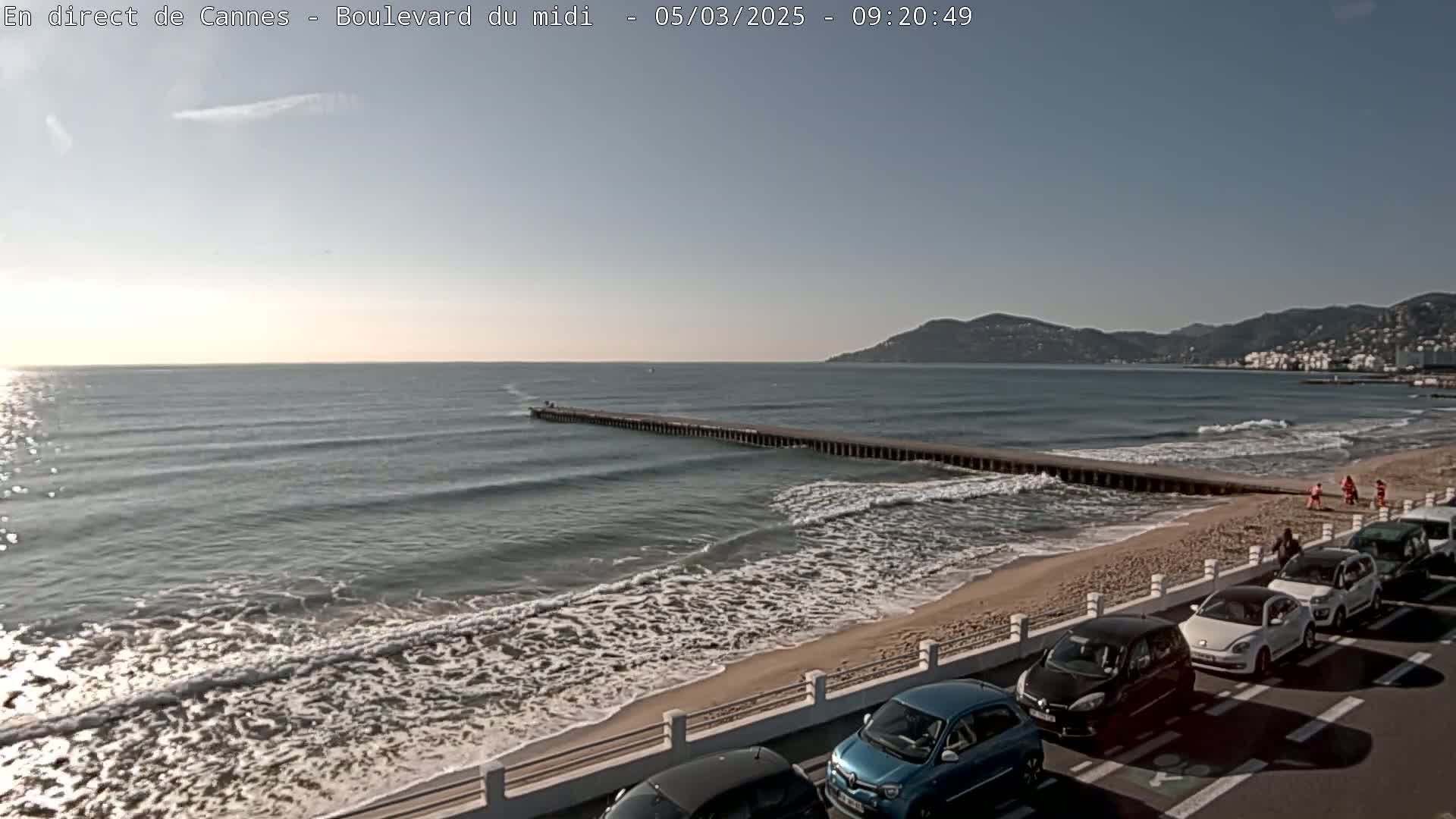 A sandy beach with gentle waves laps at the shore, under a clear sky, with cars parked along the roadside and a long pier extending into the sea.