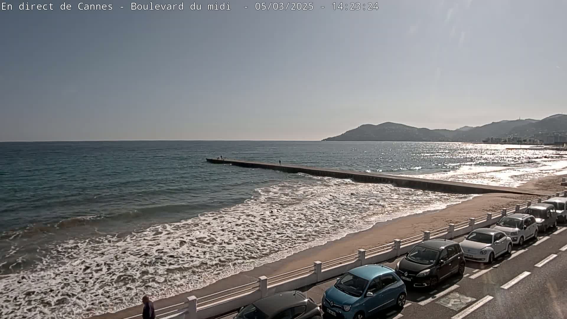 A sandy beach with gentle waves and several cars parked along a roadside next to a calm ocean under a bright, sunny sky.
