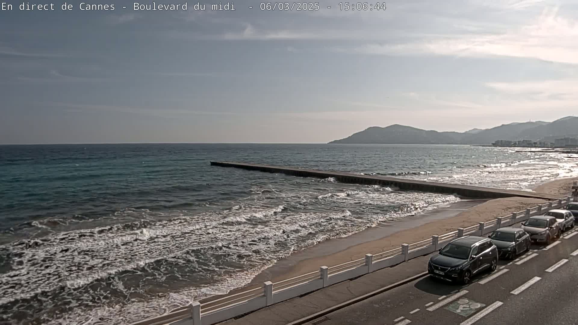A mostly sunny day shows a beach with gentle waves lapping the shore, a long pier extending into the water, and several cars parked along a road beside a white railing.