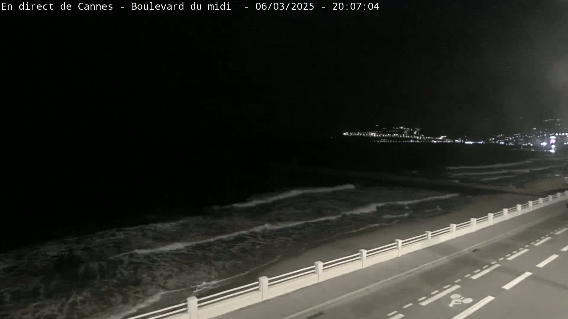 A nighttime coastal scene shows a dark ocean with small waves lapping a beach, a roadway with a low white railing separating it from the beach, and a distant city glowing with lights.