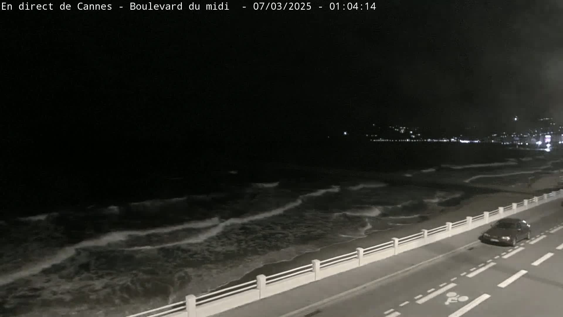 A nighttime view of a dark ocean with moderate waves crashing on a shore with a road and a low white wall beside it, a single car is visible on the road.