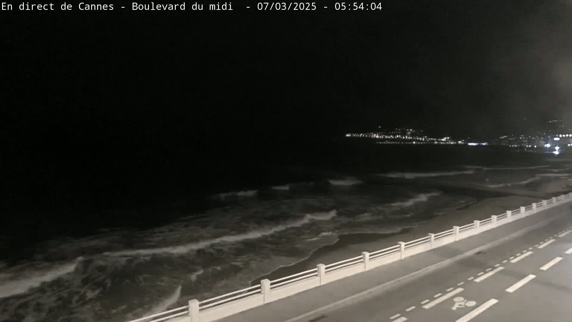 A nighttime coastal scene shows dark waves lapping a beach bordered by a roadway with a low white railing, and city lights are visible in the distance.