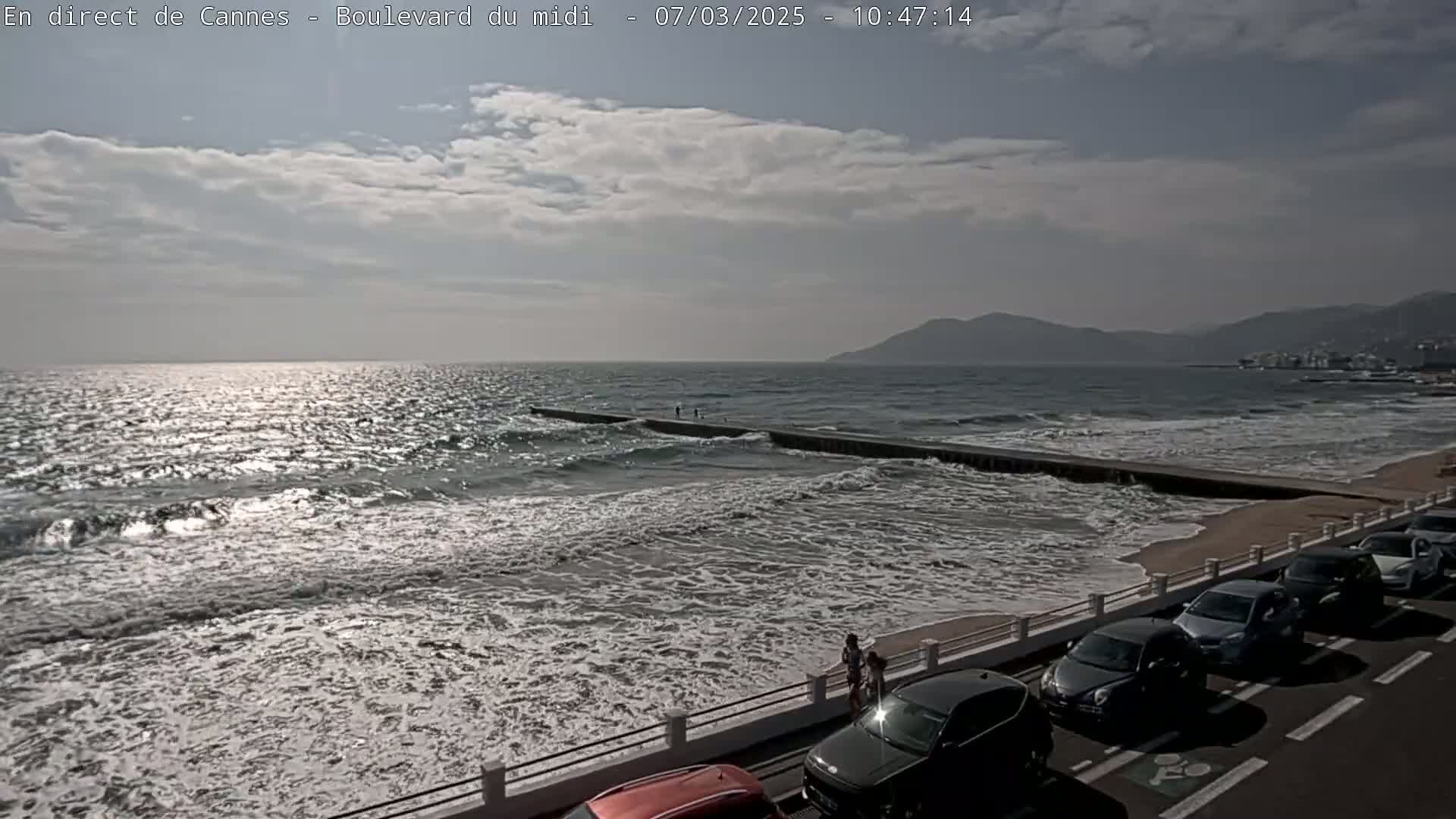 A partly cloudy day shows moderate waves breaking on a beach with a long breakwater, cars parked along a roadside, and people walking near the water.