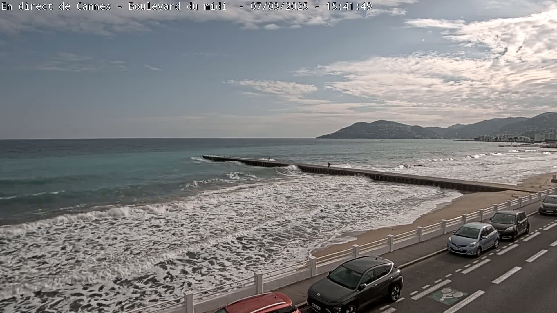 A partly cloudy day shows moderate waves breaking on a sandy beach with cars parked along a roadside next to a seawall, with mountains visible in the background.