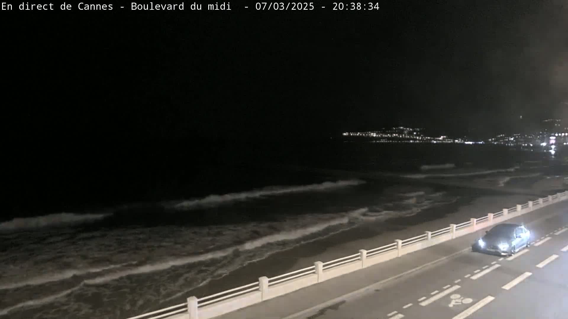 A nighttime coastal scene shows a dark ocean with waves lapping a shore, a road with a single car, and a distant city glowing with lights.