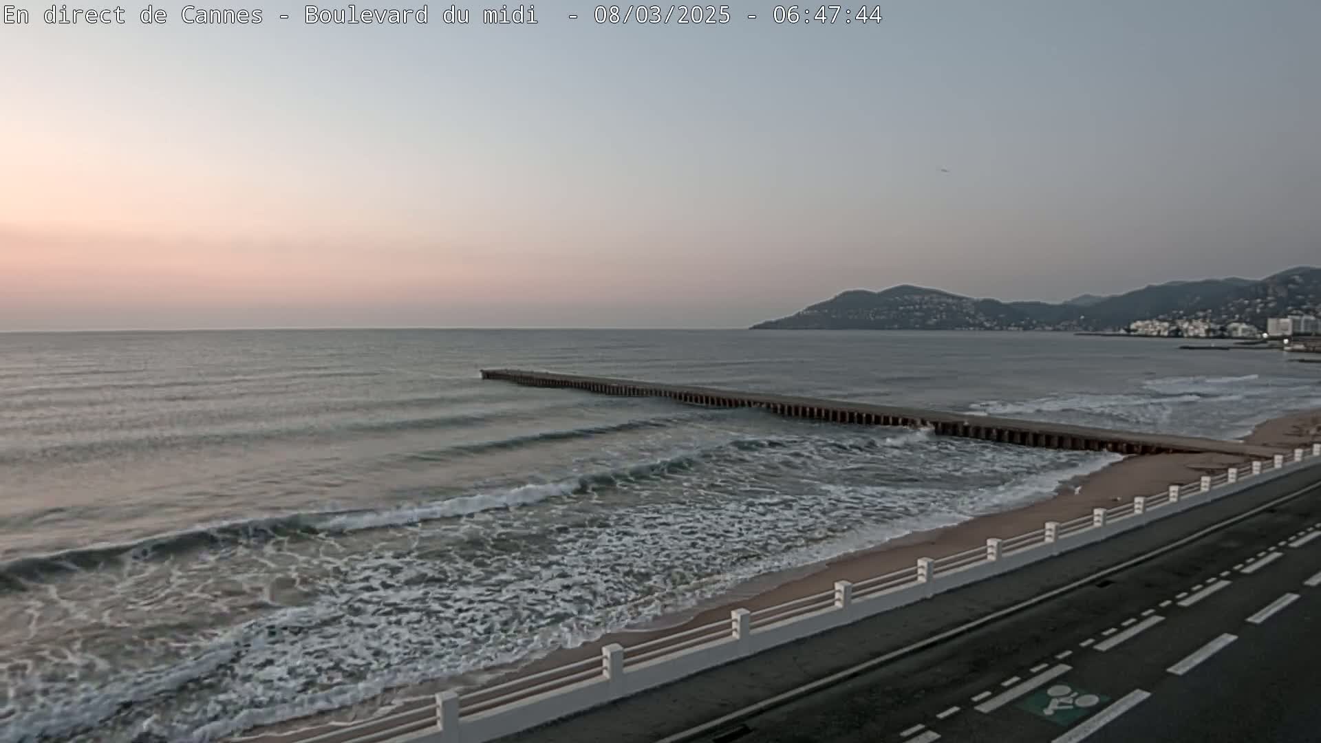 A long jetty extends into a calm sea with small waves lapping a sandy beach under a pale sunrise sky.