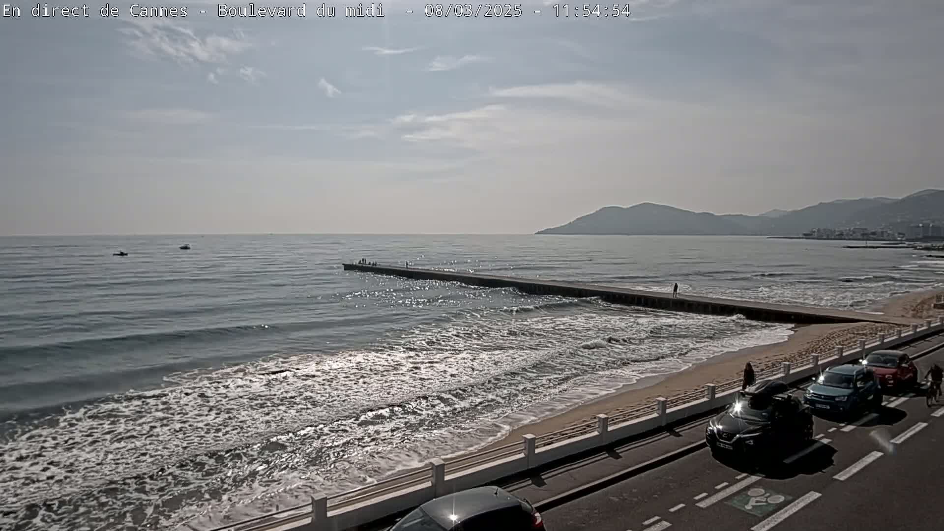 A mostly sunny day shows a beach with gentle waves, a long pier with a few people on it, and cars parked along the roadside.