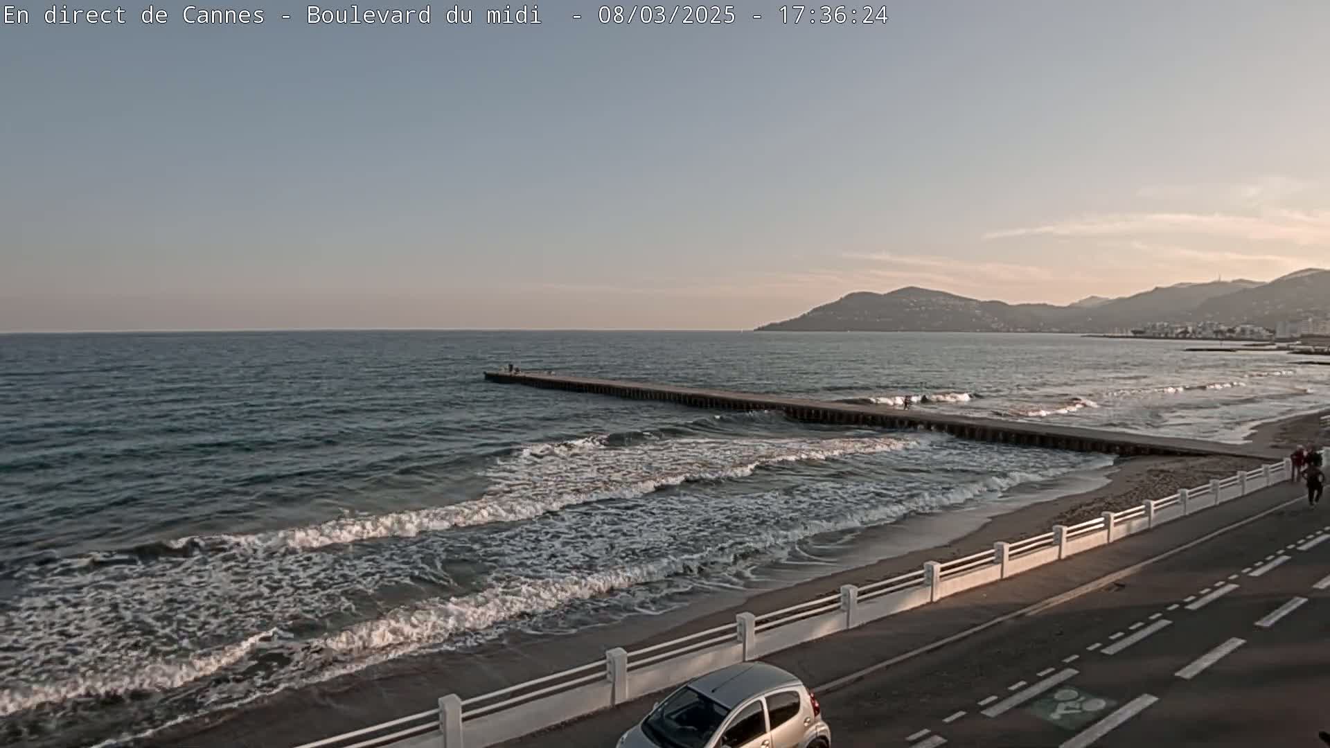 A long pier extends into a calm sea with gentle waves lapping a sandy beach under a partly cloudy, dusky sky.