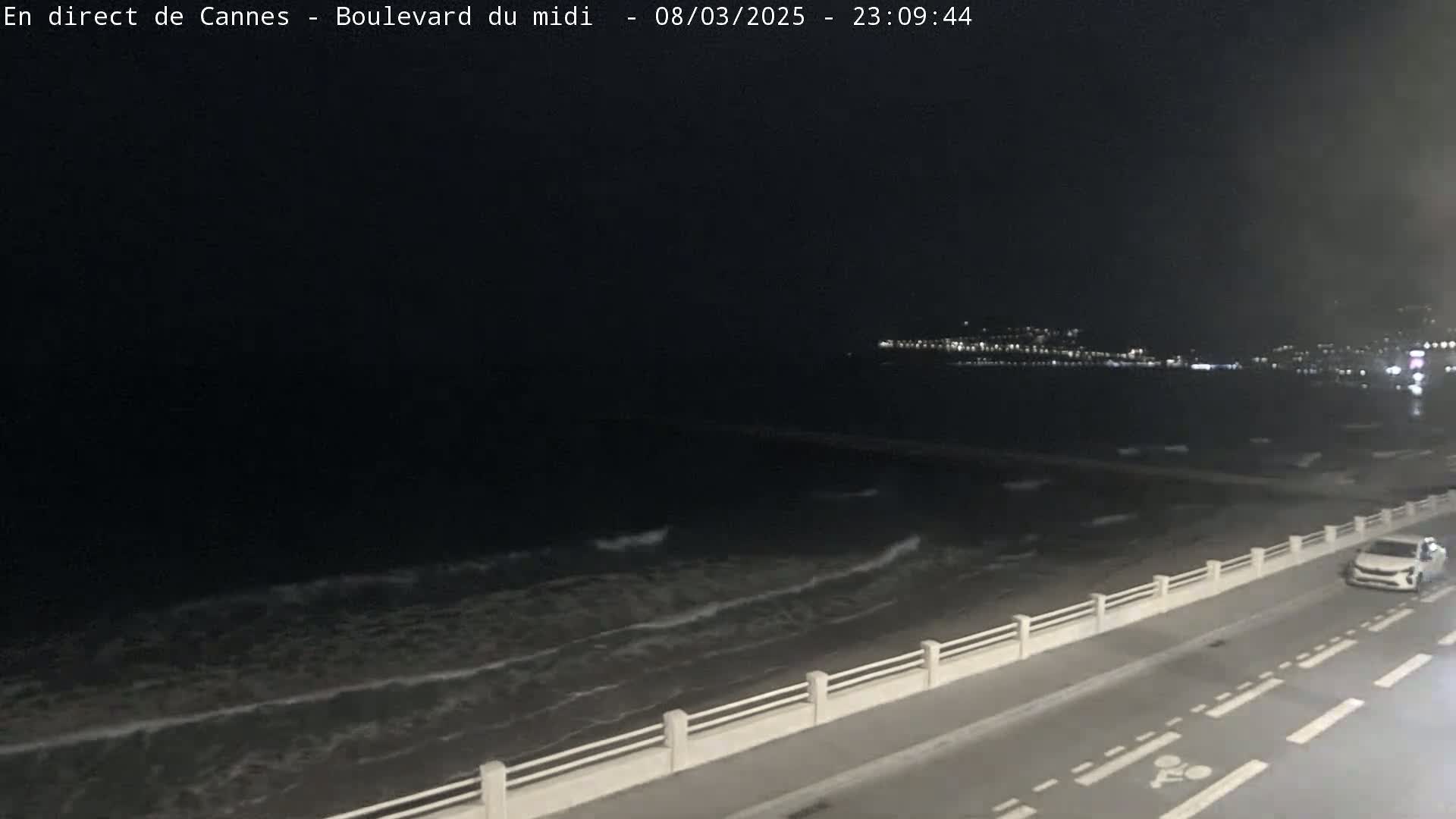 A nighttime view of a dark ocean with small waves lapping a shoreline, a road with a bike lane next to a low wall, and a single car driving on the road.