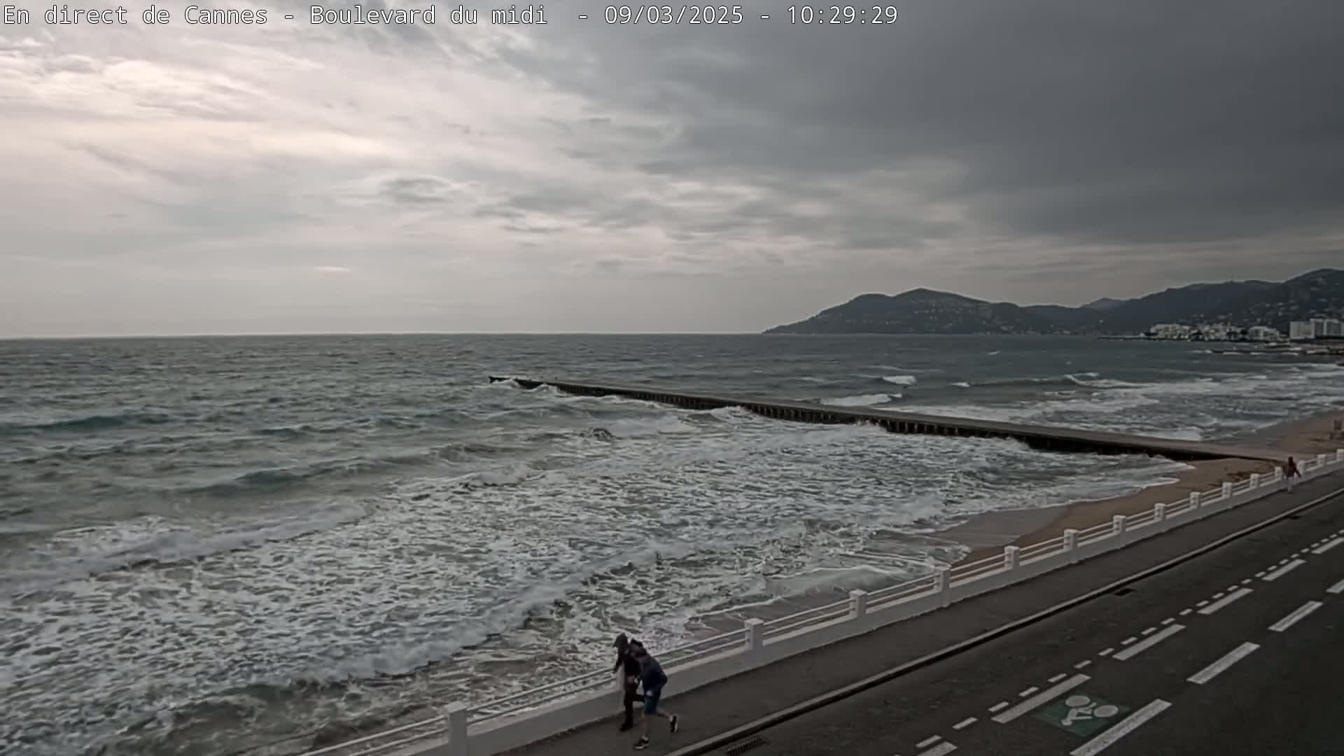 A coastal road with a white railing overlooks a choppy sea with waves breaking against a long jetty under a cloudy sky.