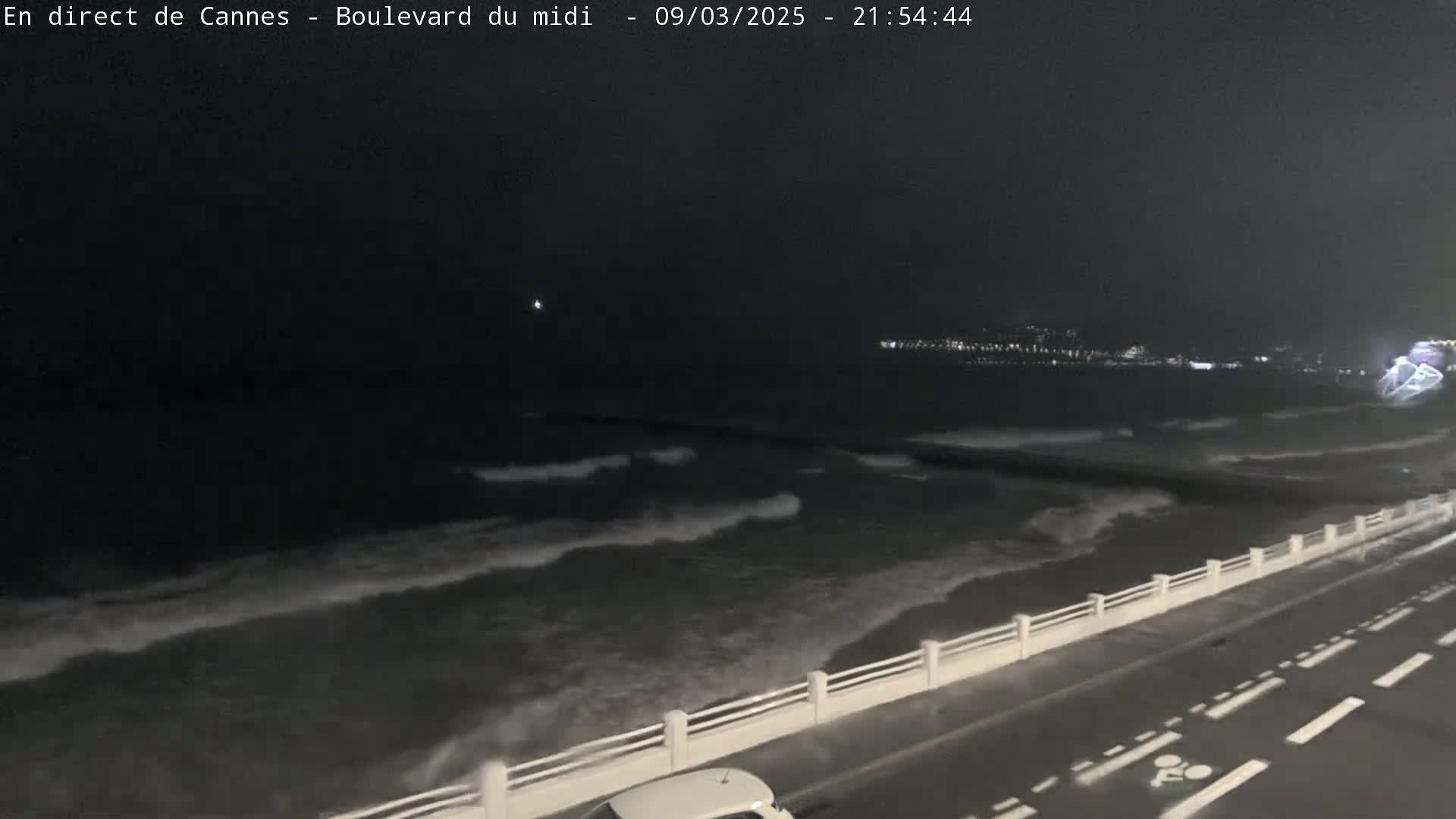 A nighttime, seaside road with a white railing separates the road from dark, wavy ocean water under a dark sky.