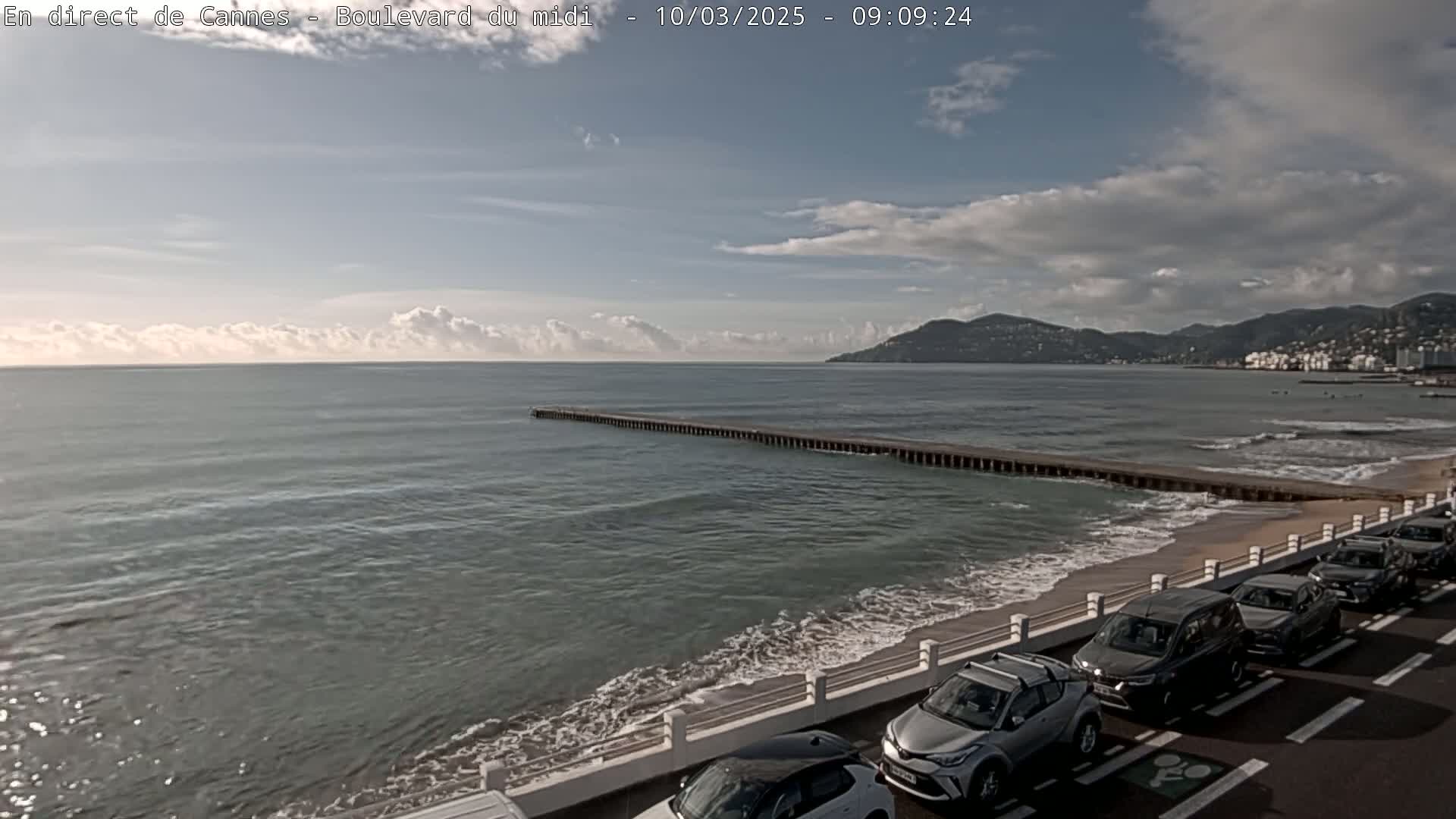 A partly cloudy day shows a beach with parked cars alongside a sea wall, overlooking a calm sea with a long pier extending into the water, and hills in the distance.