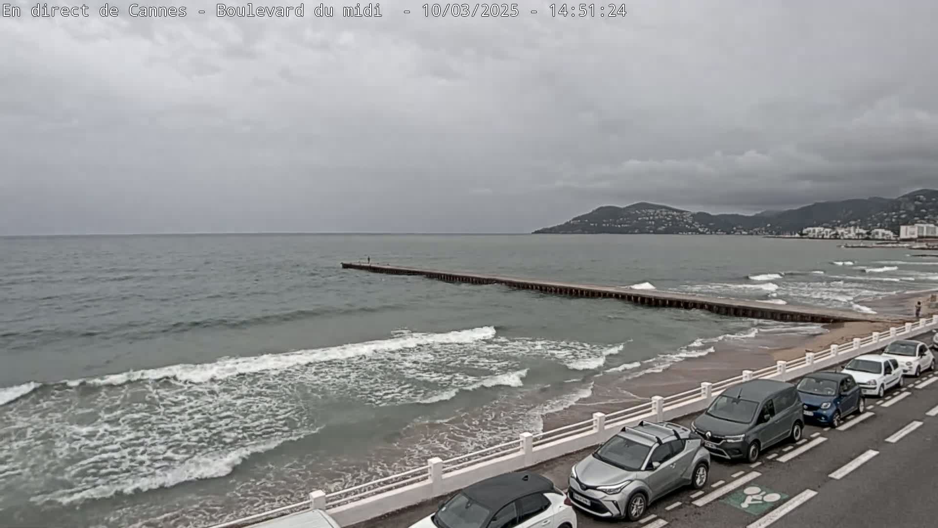A long pier extends into a wavy, gray sea under an overcast sky, with several cars parked along a beachfront road.