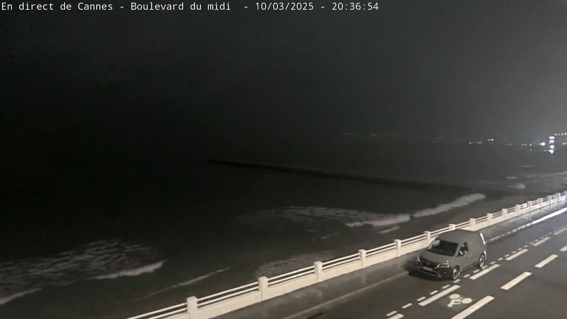 A gray van is parked on a rain-slicked road next to a sea wall at night, with dark, slightly wavy water visible beyond.