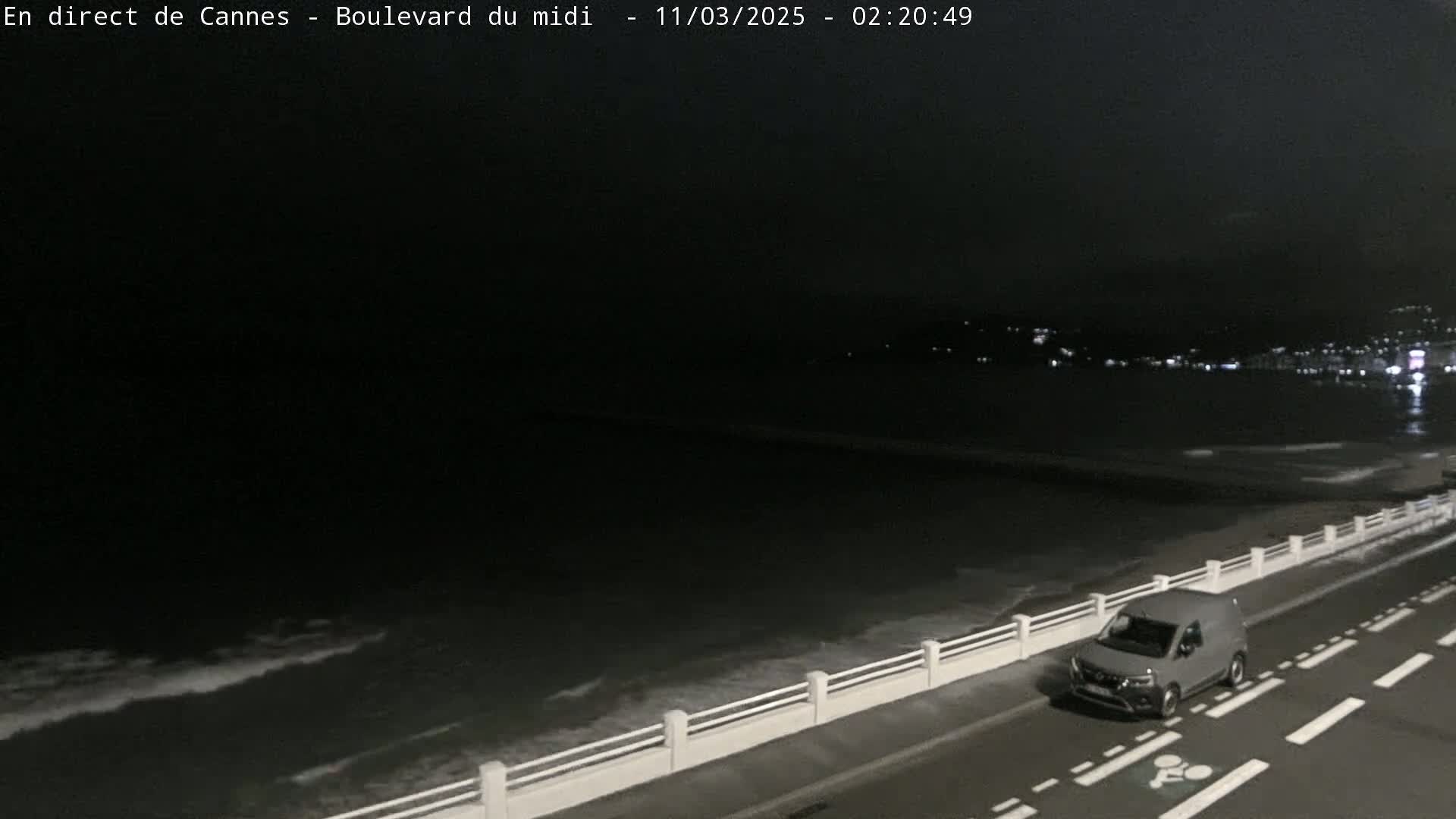 A gray van drives along a dark road beside a gently-lapping ocean at night.