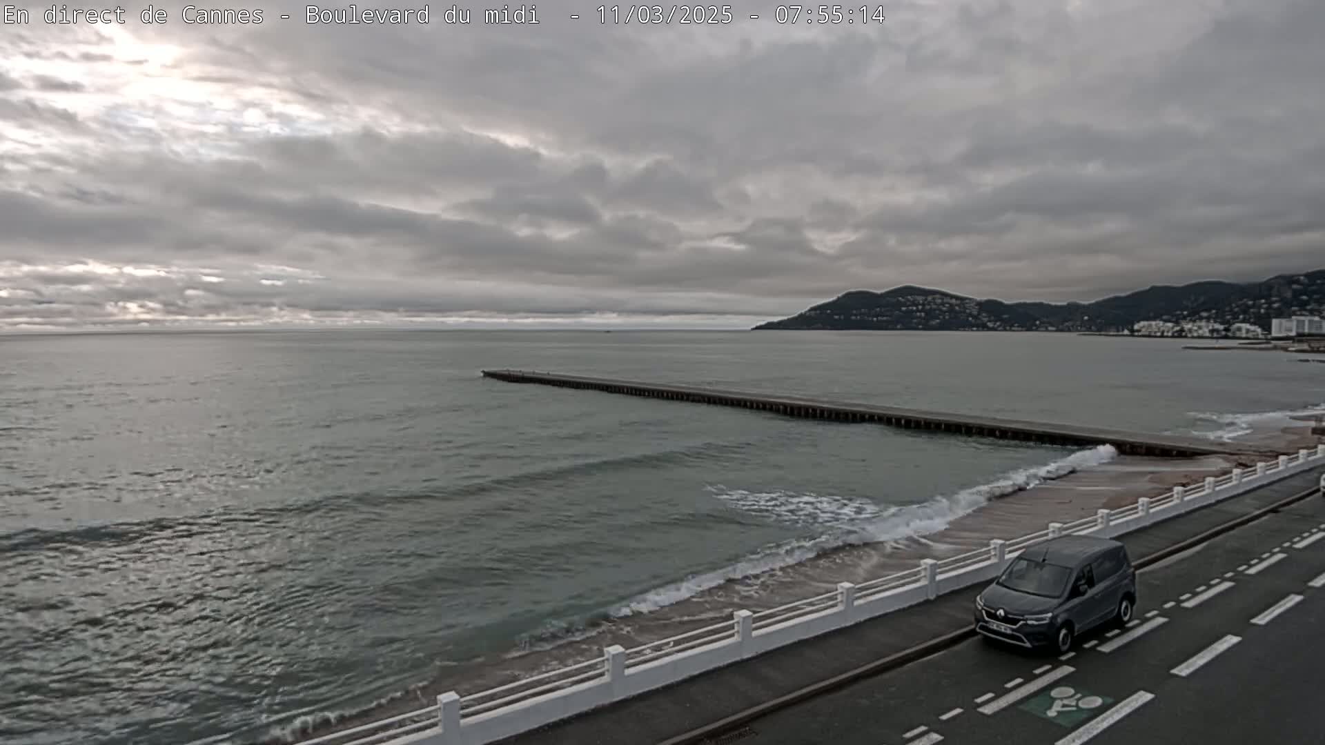 A gray minivan drives along a coastal road next to a beach with small waves under an overcast sky, a long pier extends into the gray-green sea toward a distant mountain range.