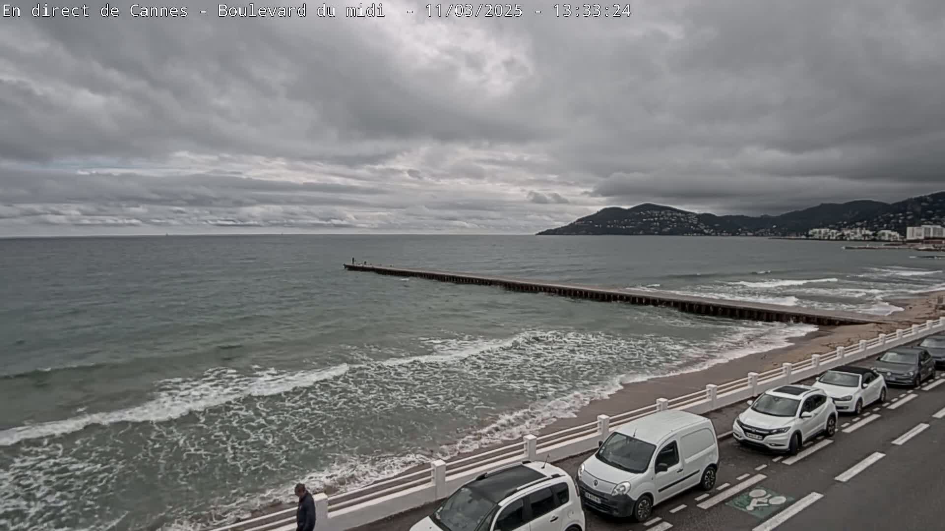 A long pier extends into a gray, wavy sea under a cloudy sky, with several cars parked along a seaside road.