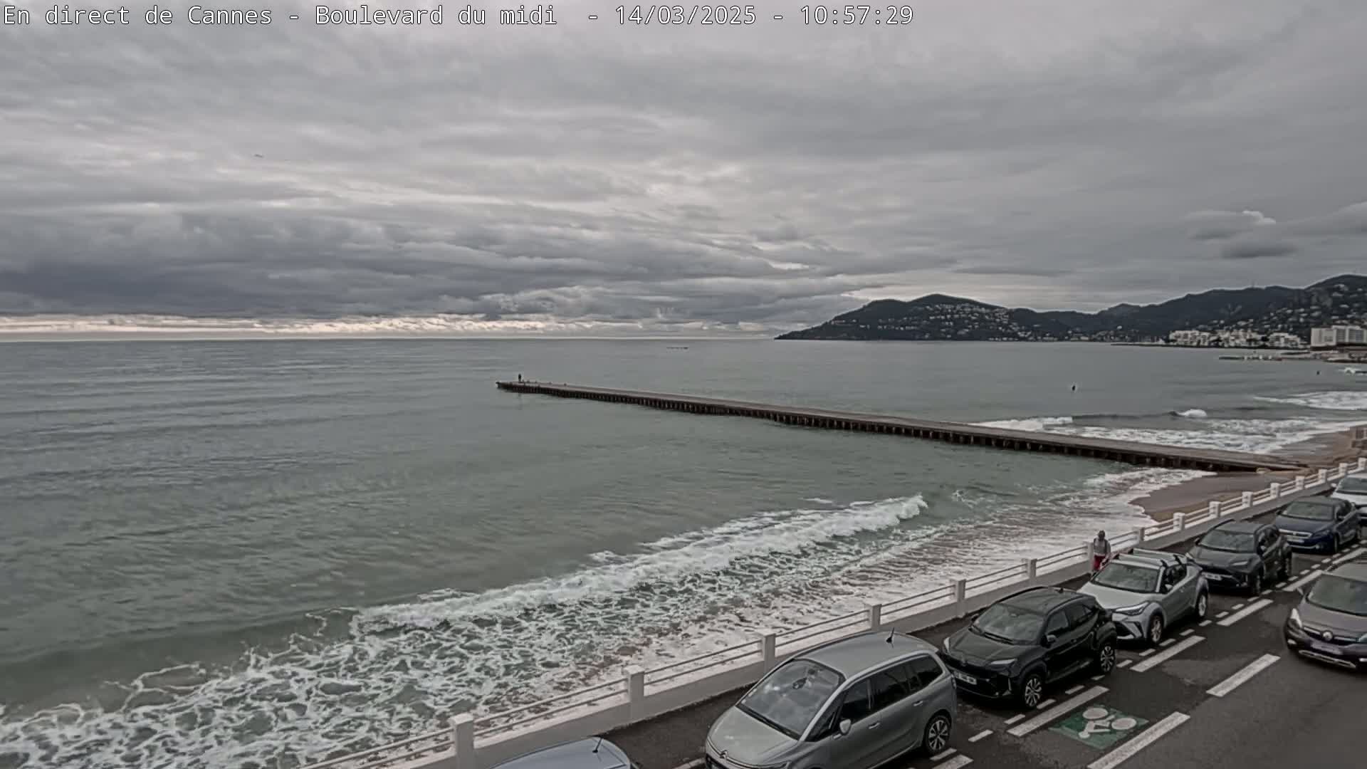 A long pier extends into a gray, somewhat choppy sea under a cloudy sky, with cars parked along a shoreline road next to a sandy beach.