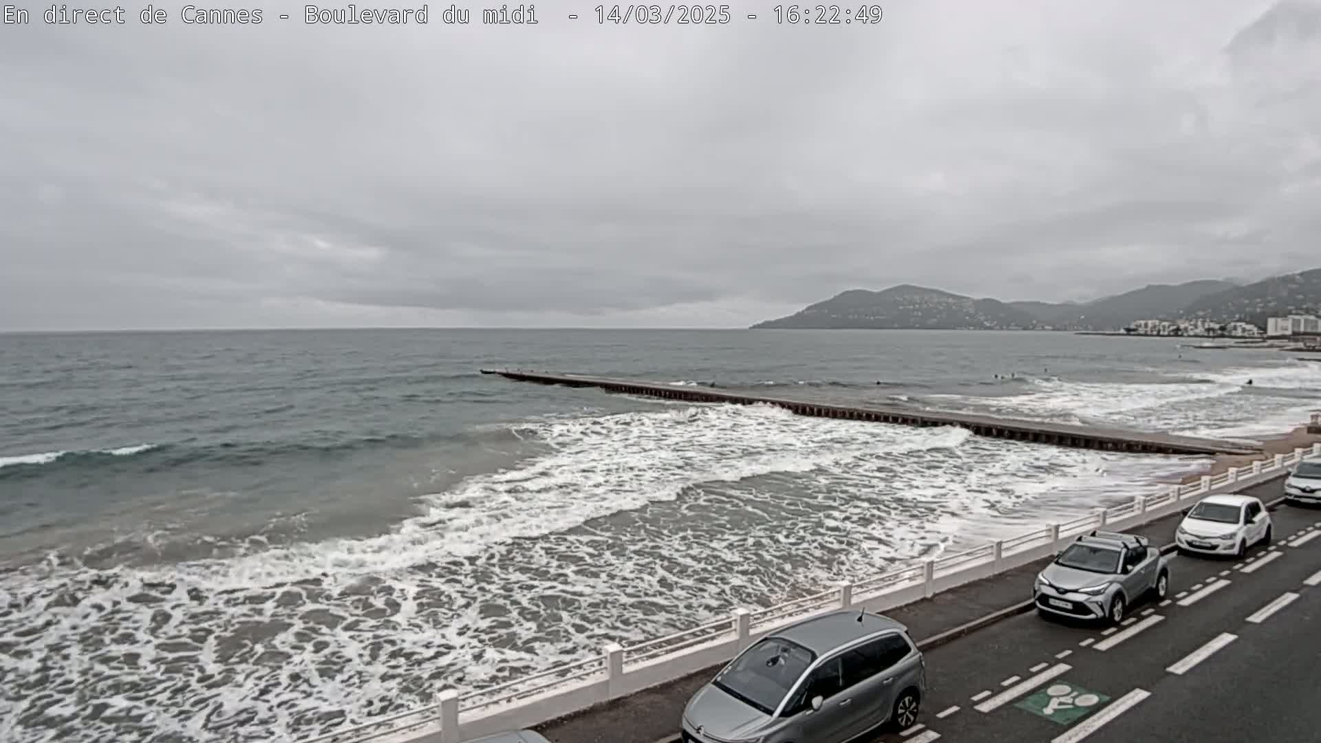 Waves crash on a beach next to a road with parked cars under an overcast sky, with distant mountains visible in the background.