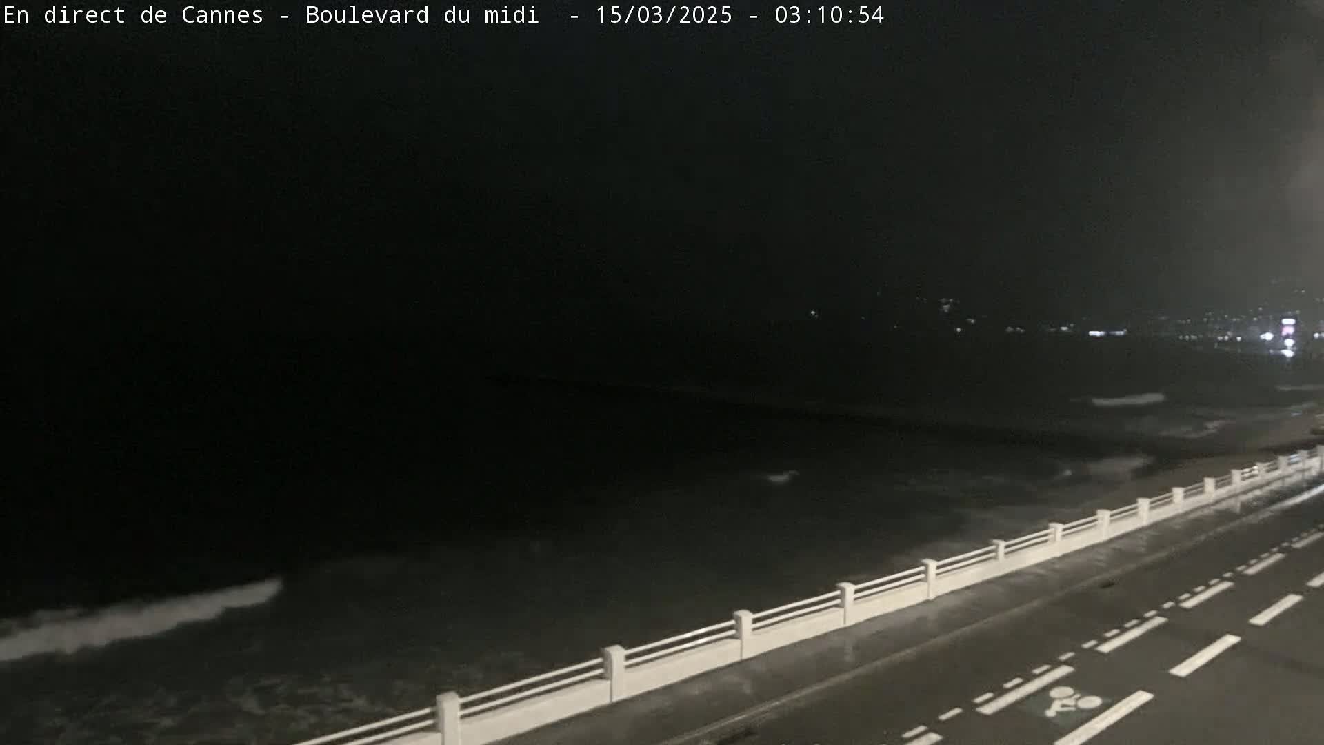 A dark, nighttime view shows a roadway next to a low seawall overlooking a choppy ocean with distant city lights.