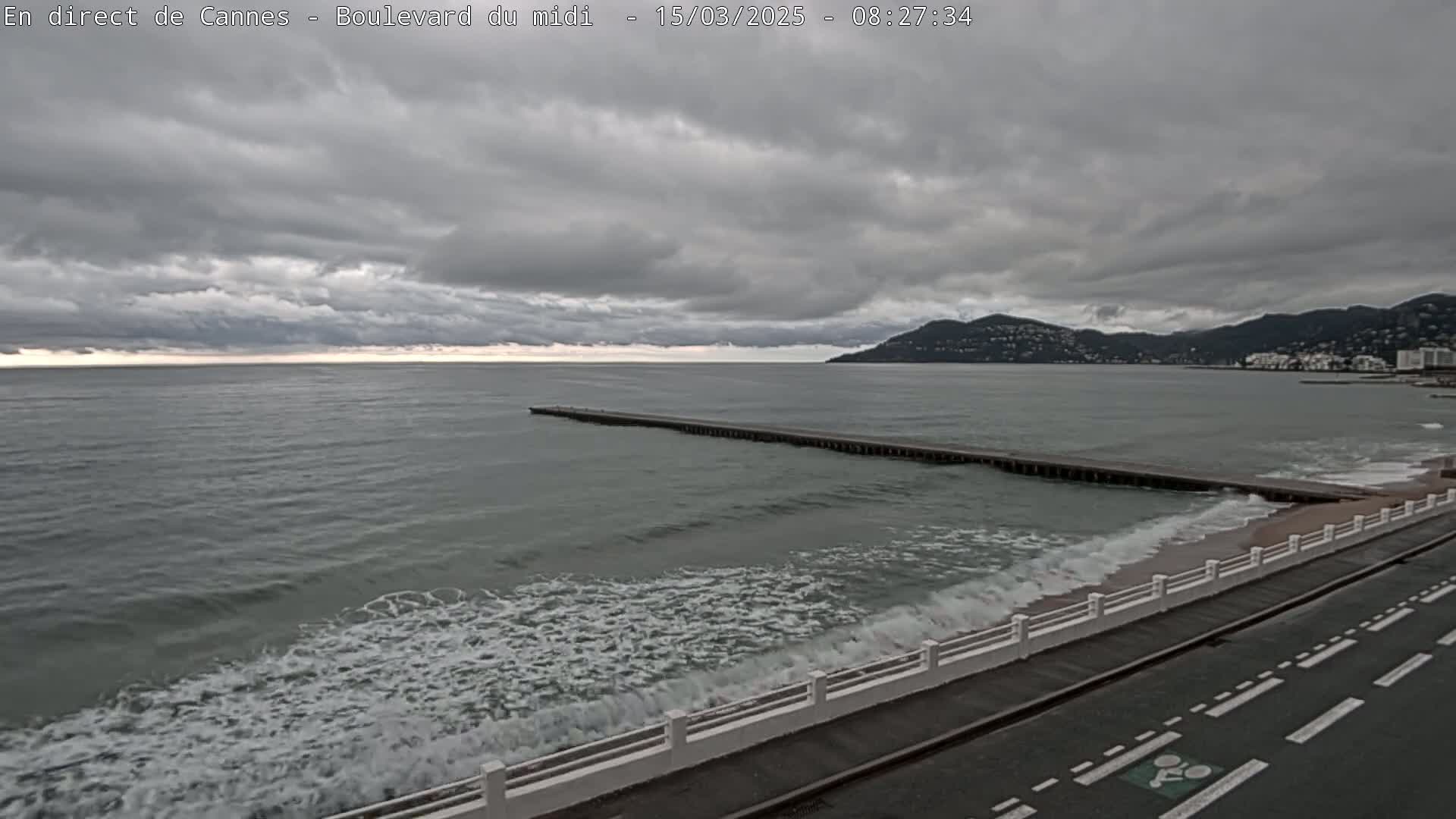 A long pier extends into a grey sea with small waves under a cloudy sky, next to a road with a white railing.
