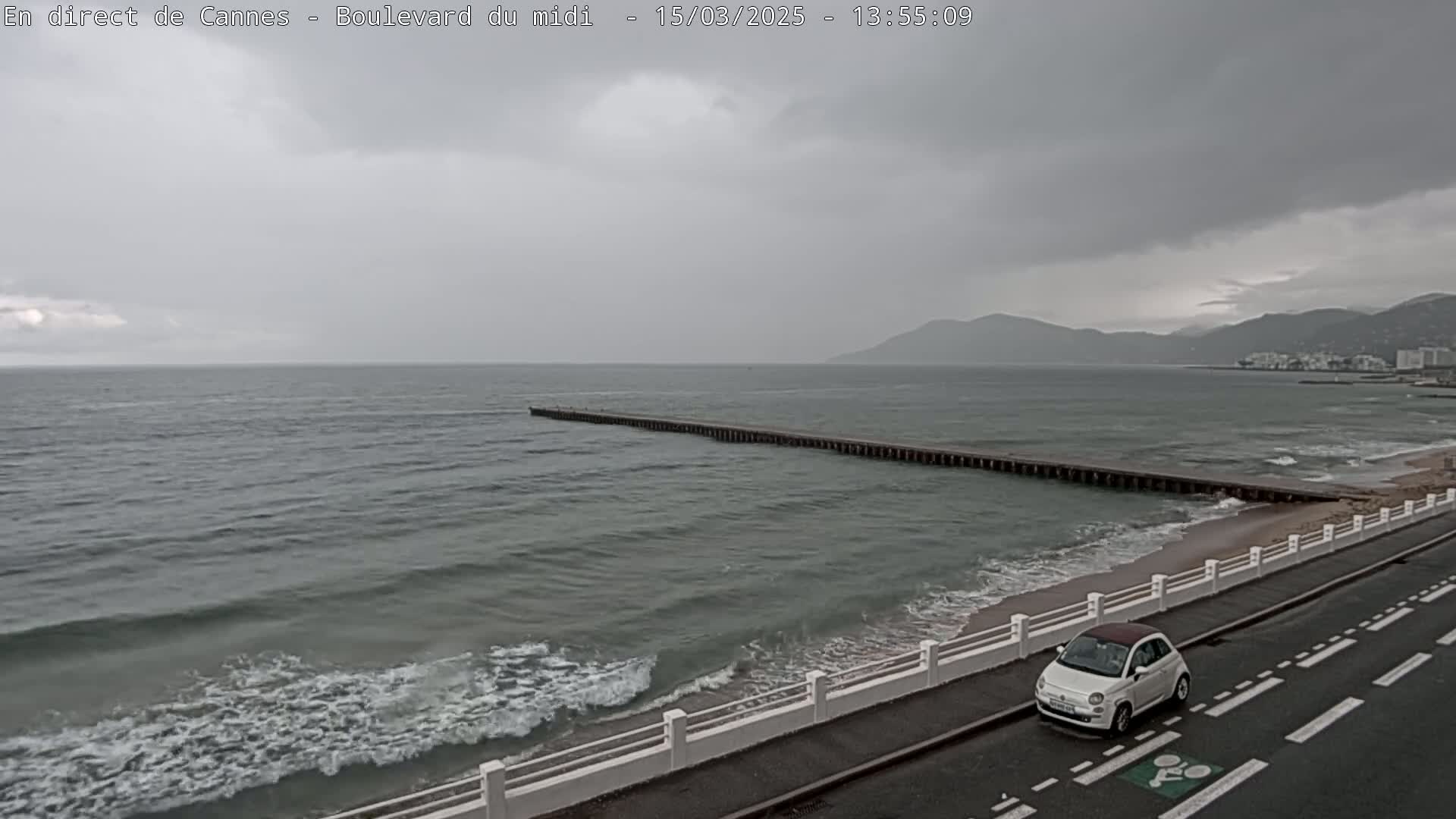 A long pier stretches into a gray, choppy sea under a cloudy sky, with a white car driving along a coastal road.