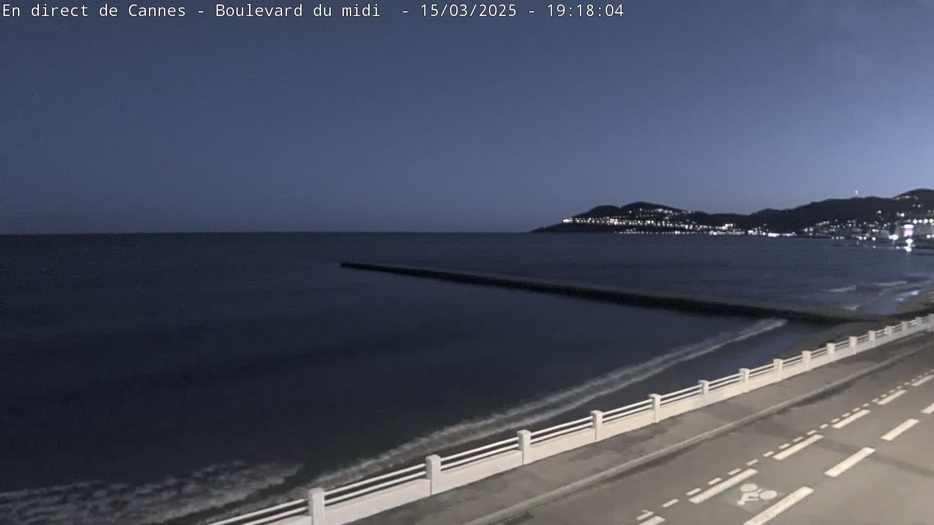 A dark-blue sea laps gently at a shoreline with a white railing and road under a twilight sky, with a town visible in the distance.