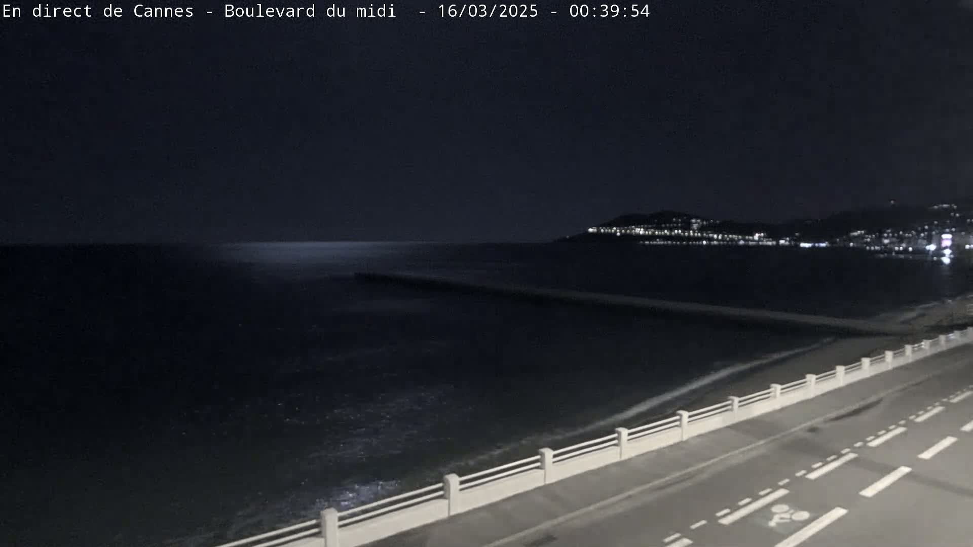 A dark, nighttime view of a calm sea lapping against a shoreline with a road and low wall, and city lights visible in the distance.