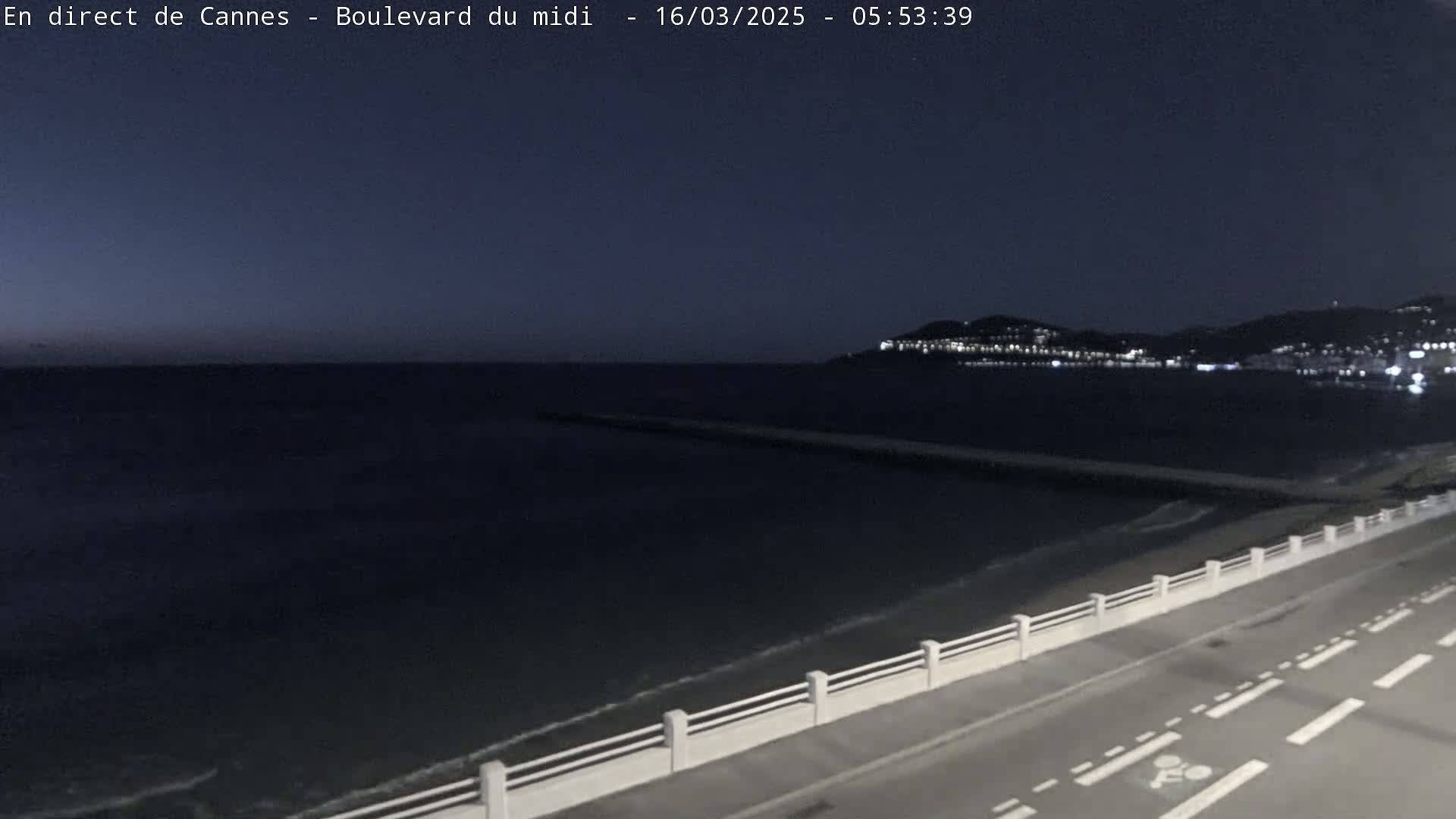 A dark, nighttime coastal scene shows a calm sea lapping gently at a shoreline road with a low white railing, beyond which are lights twinkling on a distant hillside.