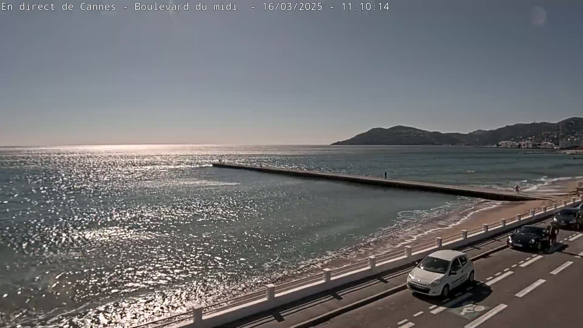 A sunny day shows a calm sea with a long jetty extending into the water, a sandy beach, and a roadway with a few cars.