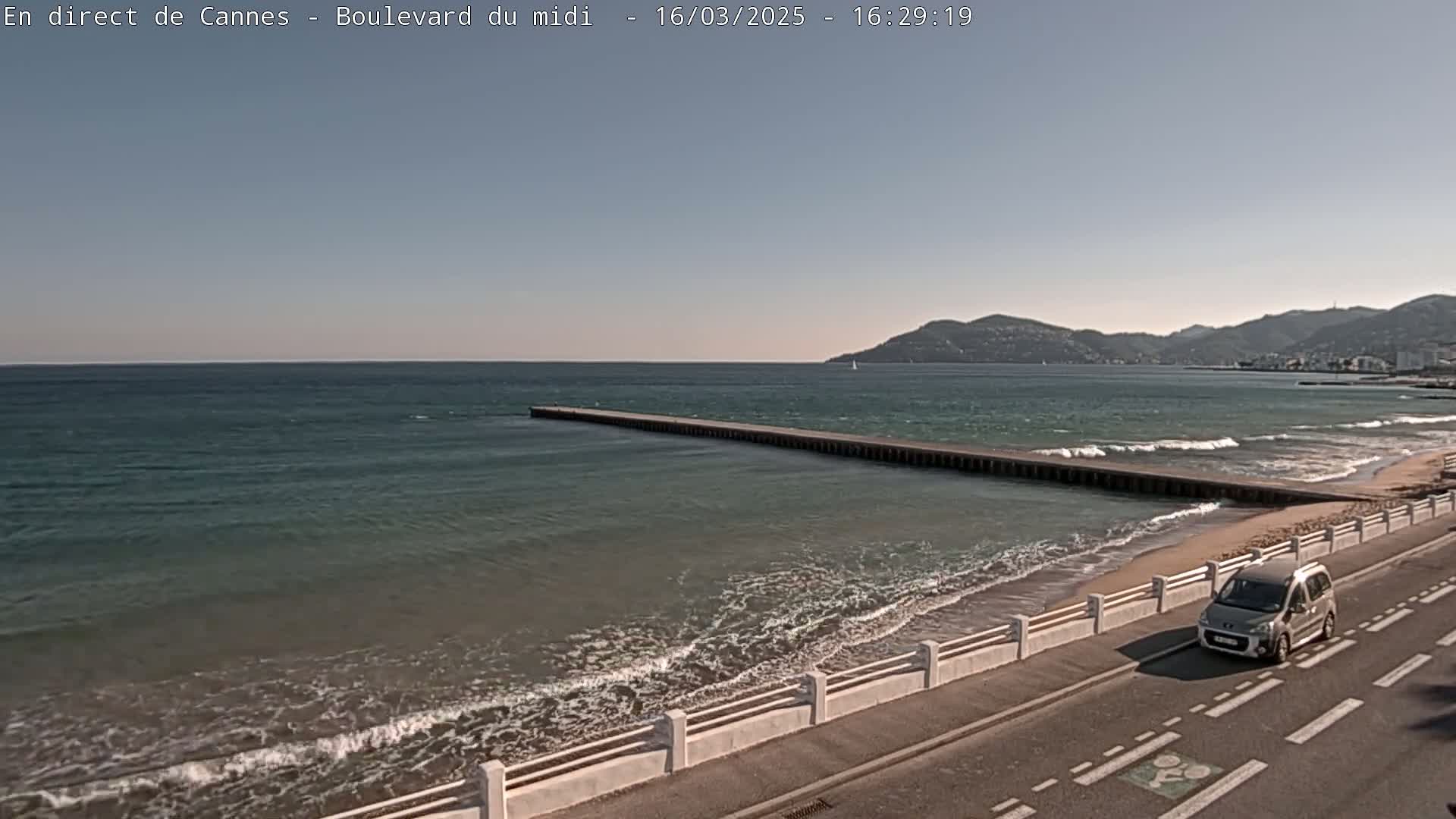 A long pier extends into a calm sea with small waves lapping a sandy beach next to a coastal road where a car is driving.