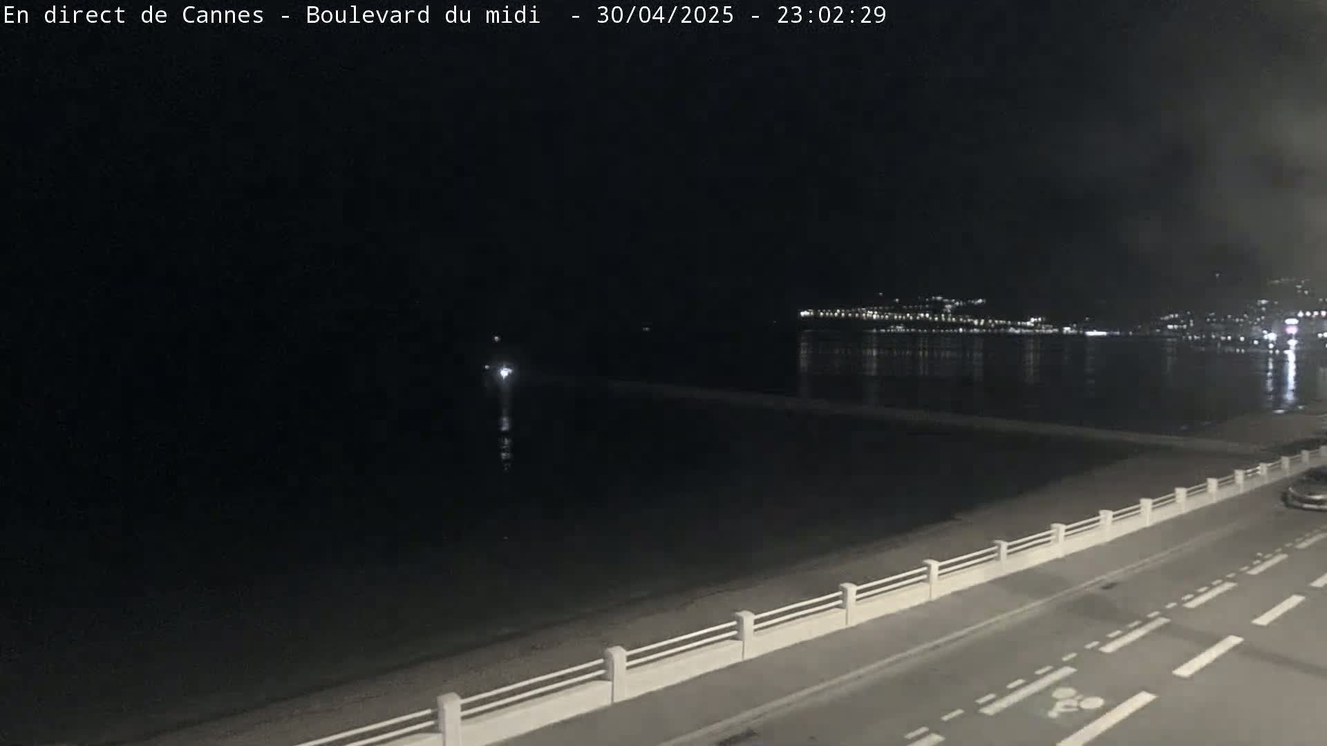 A dark nighttime scene shows a calm body of water next to a roadway with a low barrier separating it from the beach, and a distant city's lights are visible across the water.